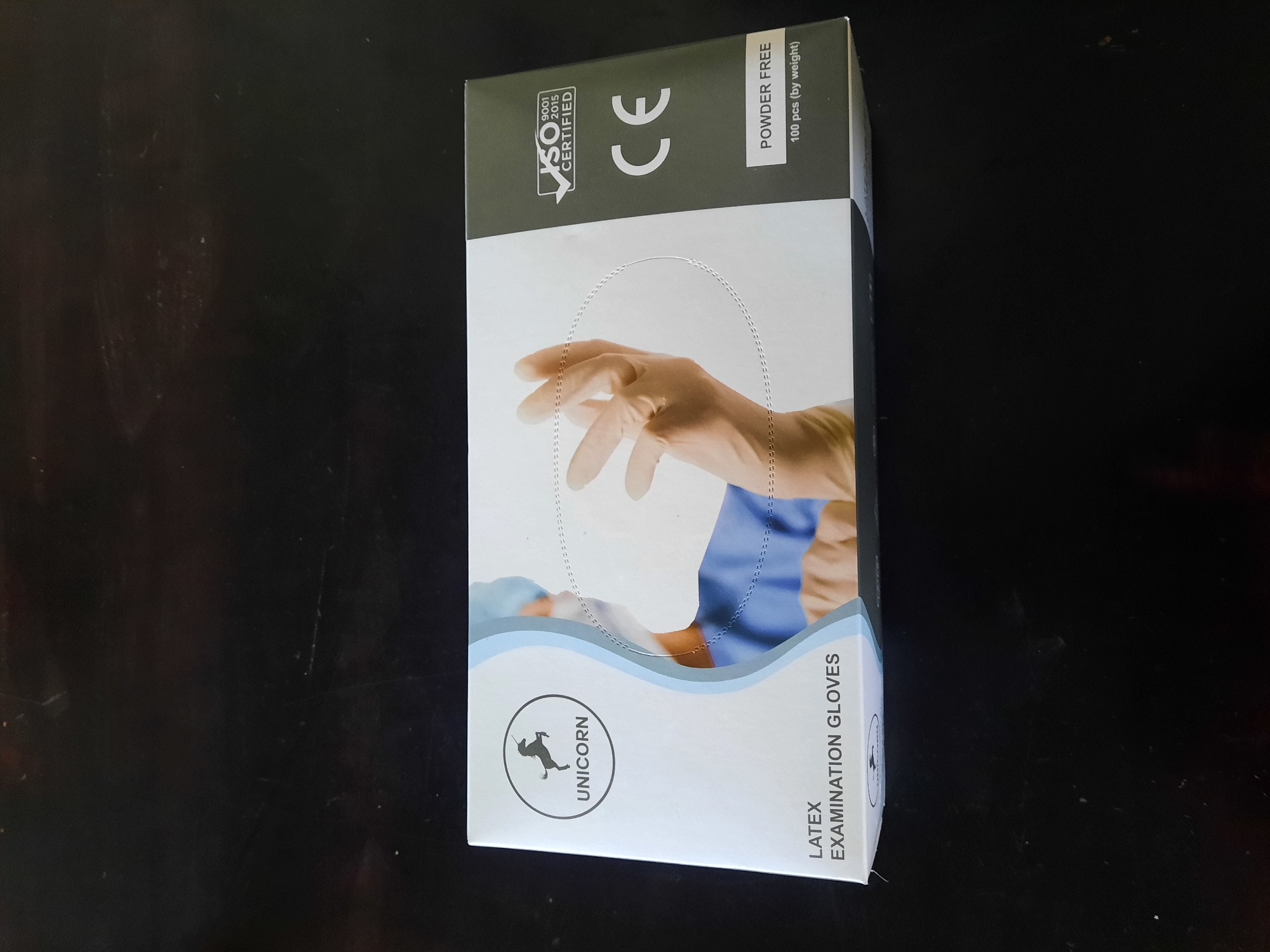A box of Unicorn brand latex examination gloves is photographed from a slightly elevated angle against a dark, textured surface. The box is predominantly white with light blue and dark grey accents. On the left side of the box, a circular logo features a silhouette of a unicorn with the word "UNICORN" below it. Below the logo, the text "LATEX EXAMINATION GLOVES" is clearly visible. The right side of the box has a dark grey background. It displays a white "CE" logo, a "ISO 9001 2015 CERTIFIED" stamp, and the text "POWDER FREE" along with "100 pcs (by weight)". A stylized image of a gloved hand and arm in a blue medical scrub is depicted on the front of the box. The background is a dark, possibly wooden, surface with subtle texture and some light reflections. There are no people or specific locations within the city of Lhokseumawe, Indonesia identifiable in this image.