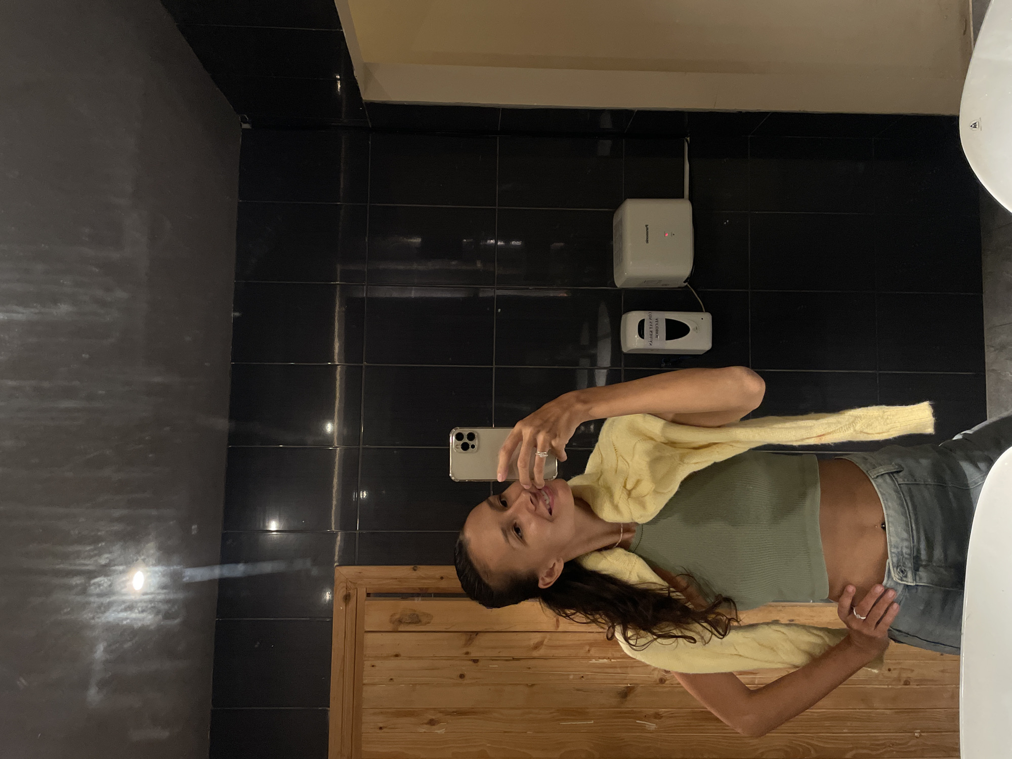 A woman is captured taking a mirror selfie in what appears to be a restroom in Phu Thuy Ward, Vietnam. The image is oriented vertically with the woman positioned in the center, holding a smartphone with her left hand to photograph her reflection. She has long dark hair, a light complexion, and wears a light green ribbed crop top, light wash jeans, and a light yellow knitted cardigan draped over her shoulders. A ring is visible on her left ring finger, and she has a belly button piercing. Her expression is a playful smile.
The background features dark, possibly black, tiled walls with visible grout lines. On the wall to the woman's right, two white devices are mounted: an upper rectangular one with a red indicator light and a "Panasonic" logo, likely a Wi-Fi router or similar electronic device, and a lower dispenser, possibly for soap or hand sanitizer, with reversed text that seems to include "WELCOME." The leftmost wall is darker, reflecting a bright artificial light source. The lower portion of the image reveals wooden paneling, suggesting a door or part of a counter. A small part of a white toilet bowl or sink is visible on the far right edge of the frame. The indoor setting is illuminated by artificial light.