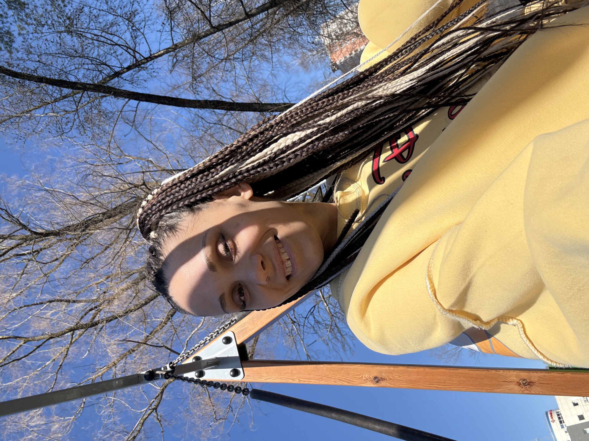 A young woman with box braids is smiling at the camera. She is wearing a yellow hoodie with a red design. The photo appears to have been taken from a low angle, looking up towards the subject and the sky. Bare tree branches are visible in the background, suggesting it may be late fall or winter. A swing set frame is visible in the bottom of the frame, partially obscuring a building. The location appears to be in an urban environment, likely Yaroslavl', Russia, based on the request.