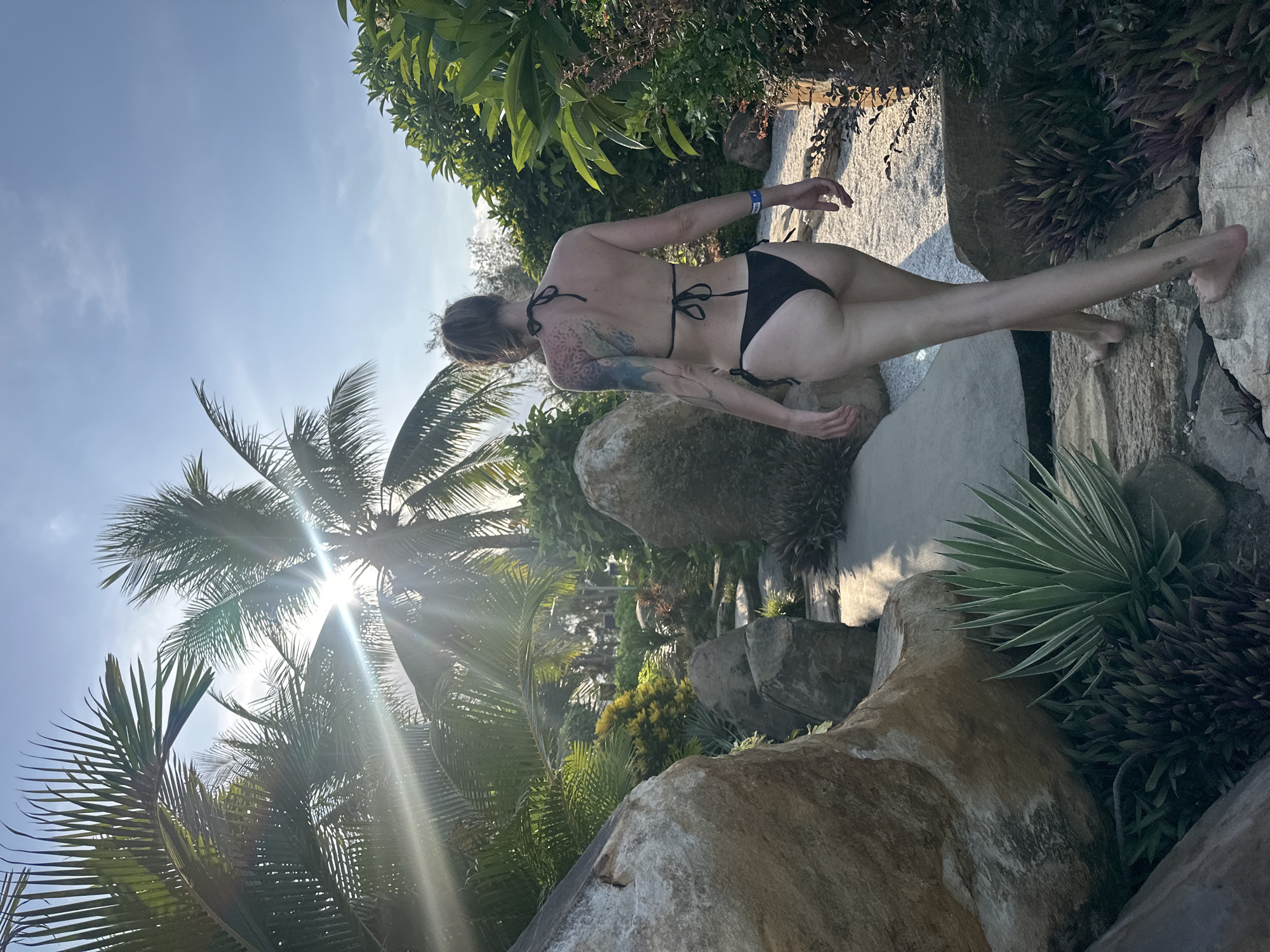 The image shows a person wearing a black bikini from the rear, standing on large rocks surrounded by lush greenery and palm trees. The sun is shining brightly, casting lens flare across the scene and creating strong shadows, suggesting it is midday. The person has tattoos on their back and arm. The tropical vegetation and the style of rocks indicate a resort or garden setting in a warm climate. The presence of palm trees and the provided location context suggest the scene is in Da Nang, Vietnam. The person is looking away from the camera, towards the sunlit foliage, and their pose is relaxed, with one arm outstretched slightly. The overall atmosphere is one of leisure and enjoyment in a natural, sun-drenched environment. No visible text is present in the image.