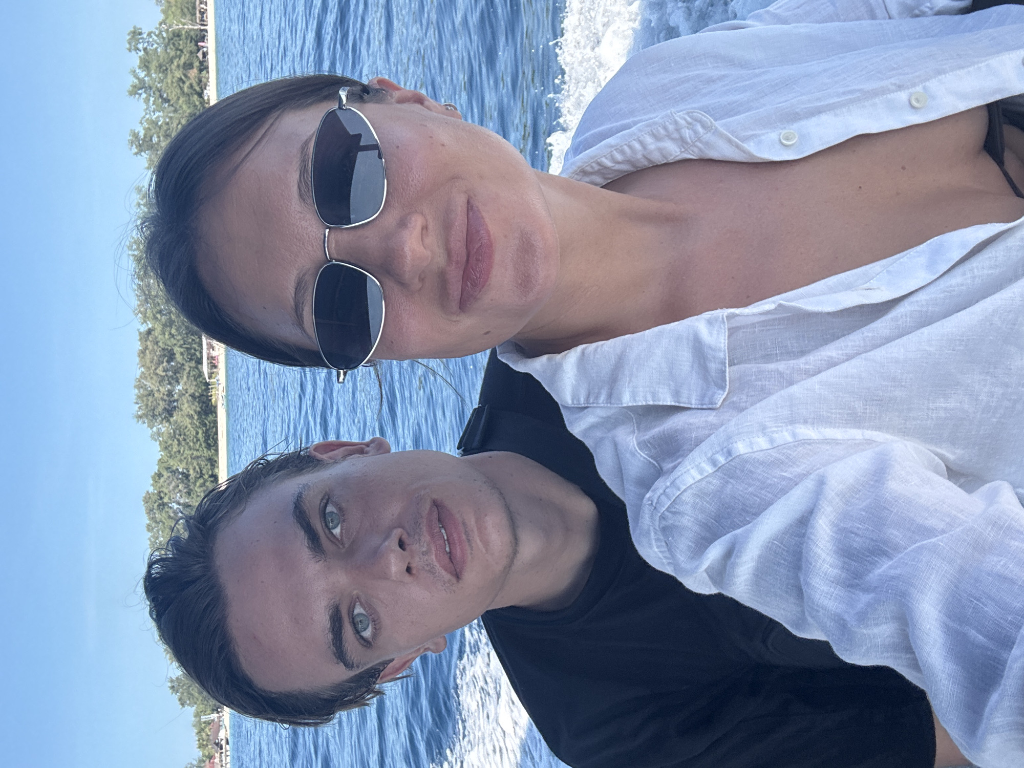 Two individuals are positioned on a boat moving across water. A woman, wearing sunglasses and a white collared shirt, is positioned higher in the frame. A man with blue eyes, wearing a black shirt, is beneath her. The water is a deep blue with white wake from the boat's movement visible. The background shows a coastline with trees under a blue sky. The city is Unknown, Indonesia.