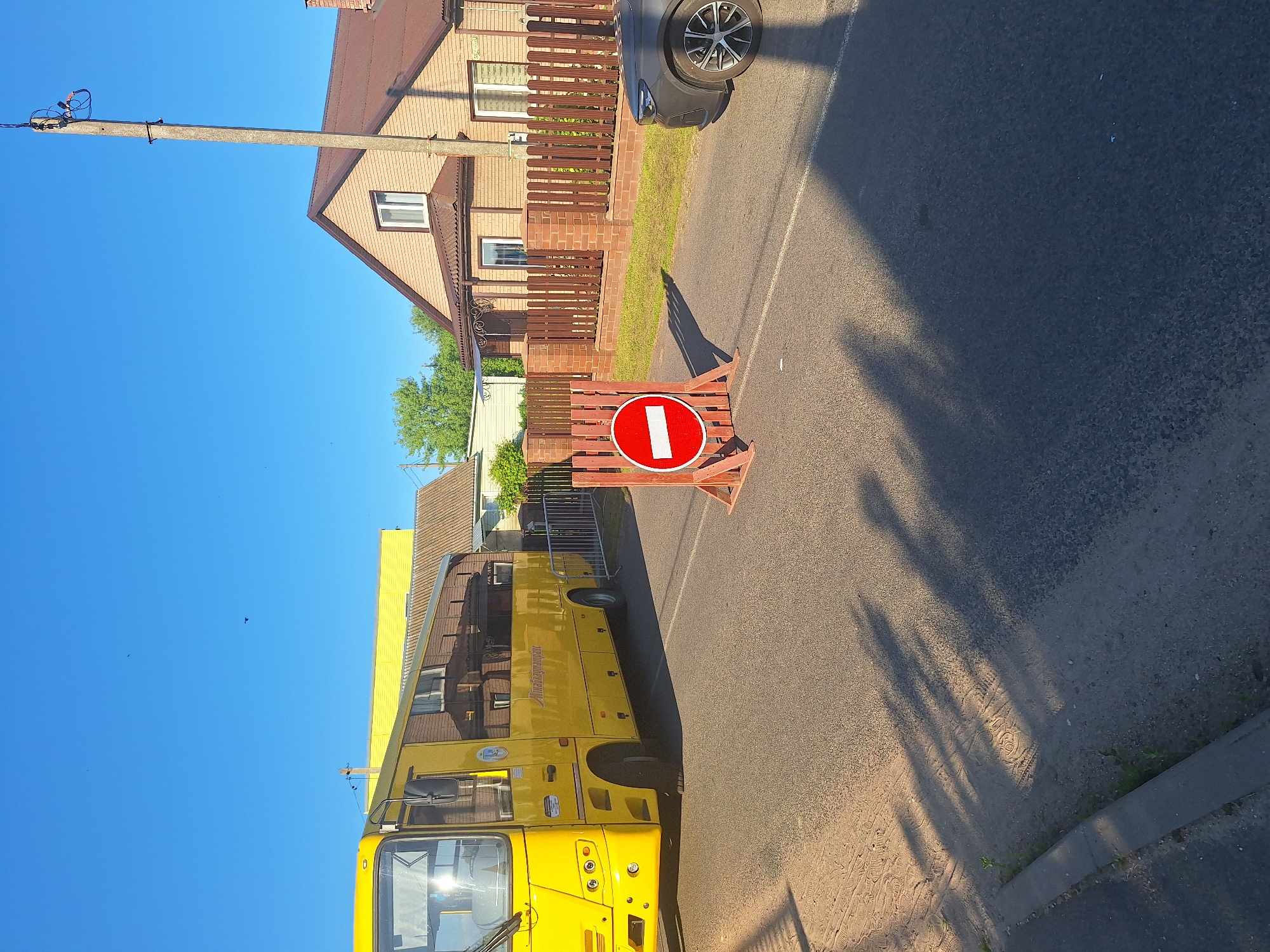 In the village of Zimkovichi, within the Zimkovichi rural council of Belarus, a bright yellow bus is parked on a paved road. To the right of the bus, a temporary wooden barrier with a red and white "do not enter" sign is placed on the road, indicating a closure. A dark-colored car is parked to the right of the barrier. The background features residential houses with brick facades and brown roofs, surrounded by wooden fences. Green trees and clear blue sky complete the scene. The lighting suggests it is daytime.