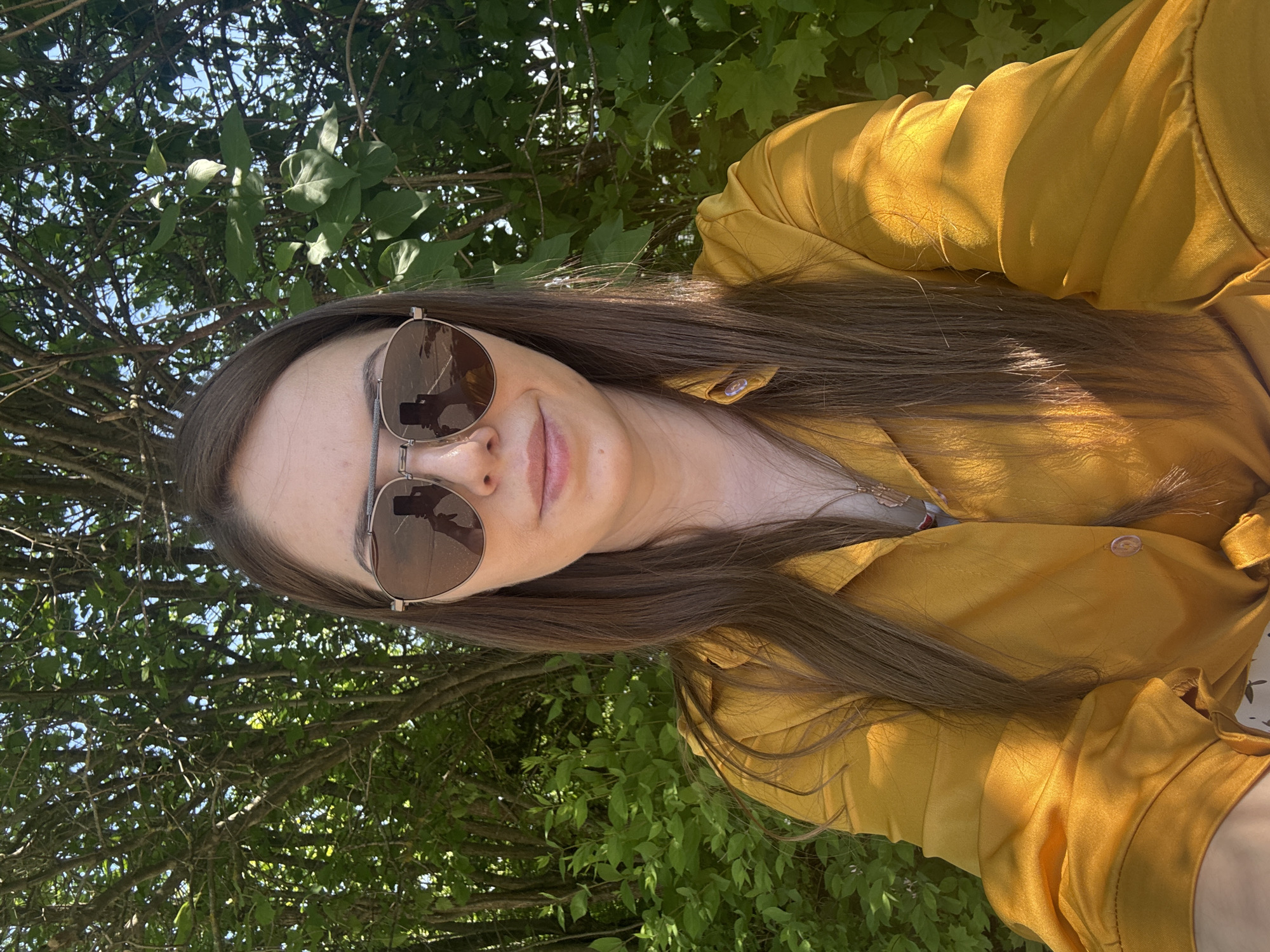 The image shows a person with long brown hair wearing sunglasses and a yellow collared shirt. The person is outdoors, with greenery and trees visible in the background. The scene appears to be taken during the day. The location is not determinable from the image.