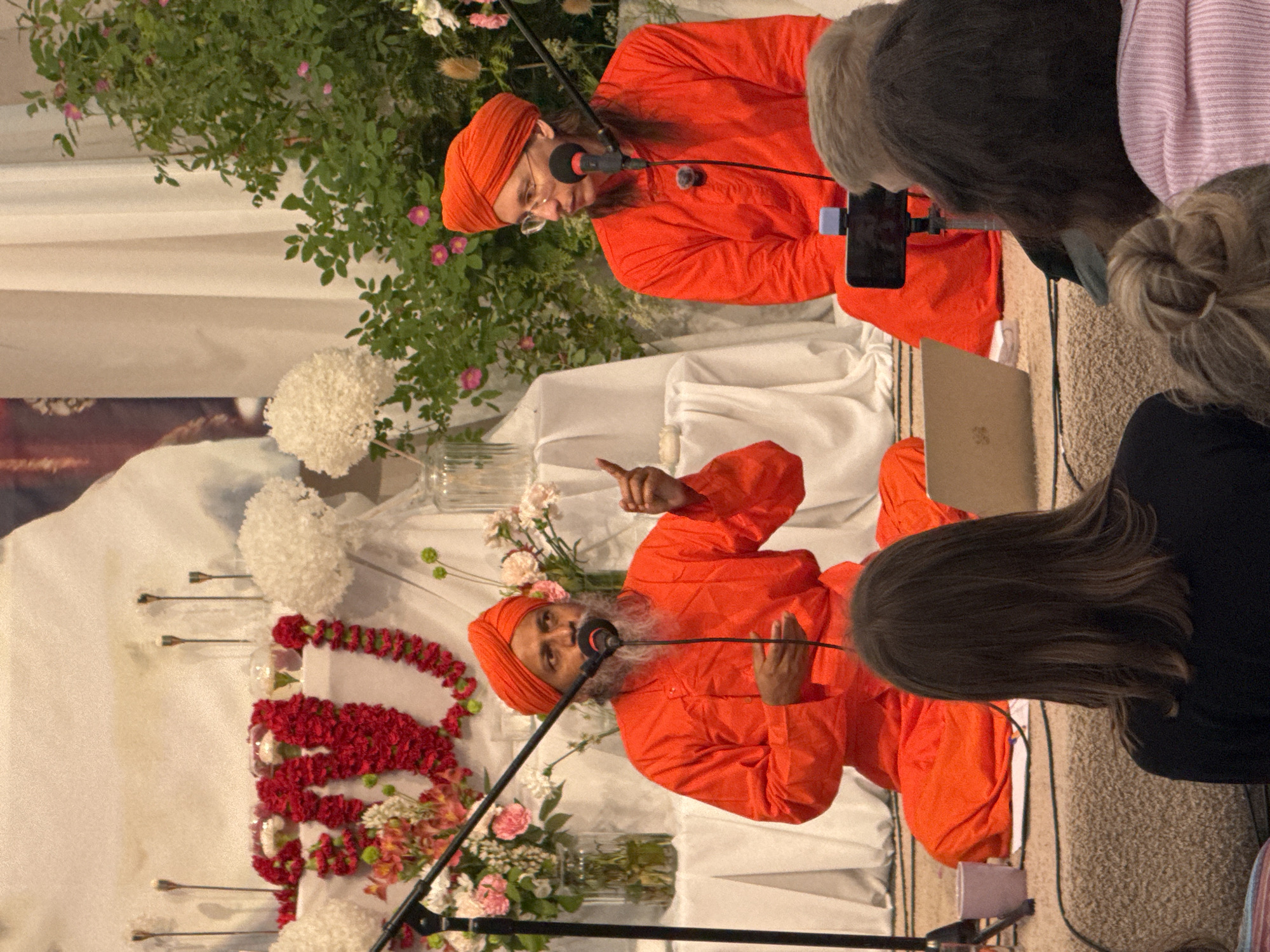 The image shows two men wearing orange turbans and robes, seated on a carpeted platform. The man on the left has a beard and is speaking into a microphone, gesturing with his right hand. The man on the right is wearing glasses and looking to his left. Behind them, there is a white backdrop decorated with red flower garlands, white floral arrangements, and two clear vases containing flowers. To the right of the men, a lush green bush with pink flowers is visible. In the foreground, several people are seated, their backs to the camera, with a laptop and a smartphone on a tripod positioned on the platform. The scene appears to be a religious or spiritual gathering. The location in Zhirnosovo, Russia, is not discernable from the image itself.