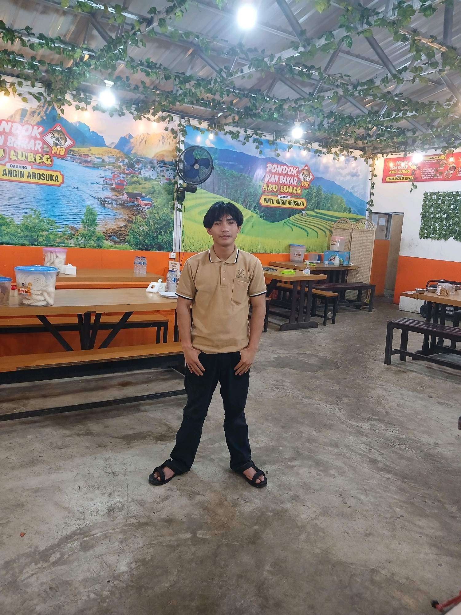 The image depicts an indoor setting, likely a restaurant or food stall, with a man standing in the center. The man is wearing a tan polo shirt and black pants. He is facing the camera with his hands in his pockets. In the background, there are wooden tables and benches. A large mural depicting a scenic landscape with mountains and a body of water adorns the back wall. The ceiling is covered with artificial greenery. The floor is concrete. The location is identified as Koto Gaek Guguak, Indonesia, though no specific landmarks within the city are visible. The overall atmosphere appears casual and relaxed.