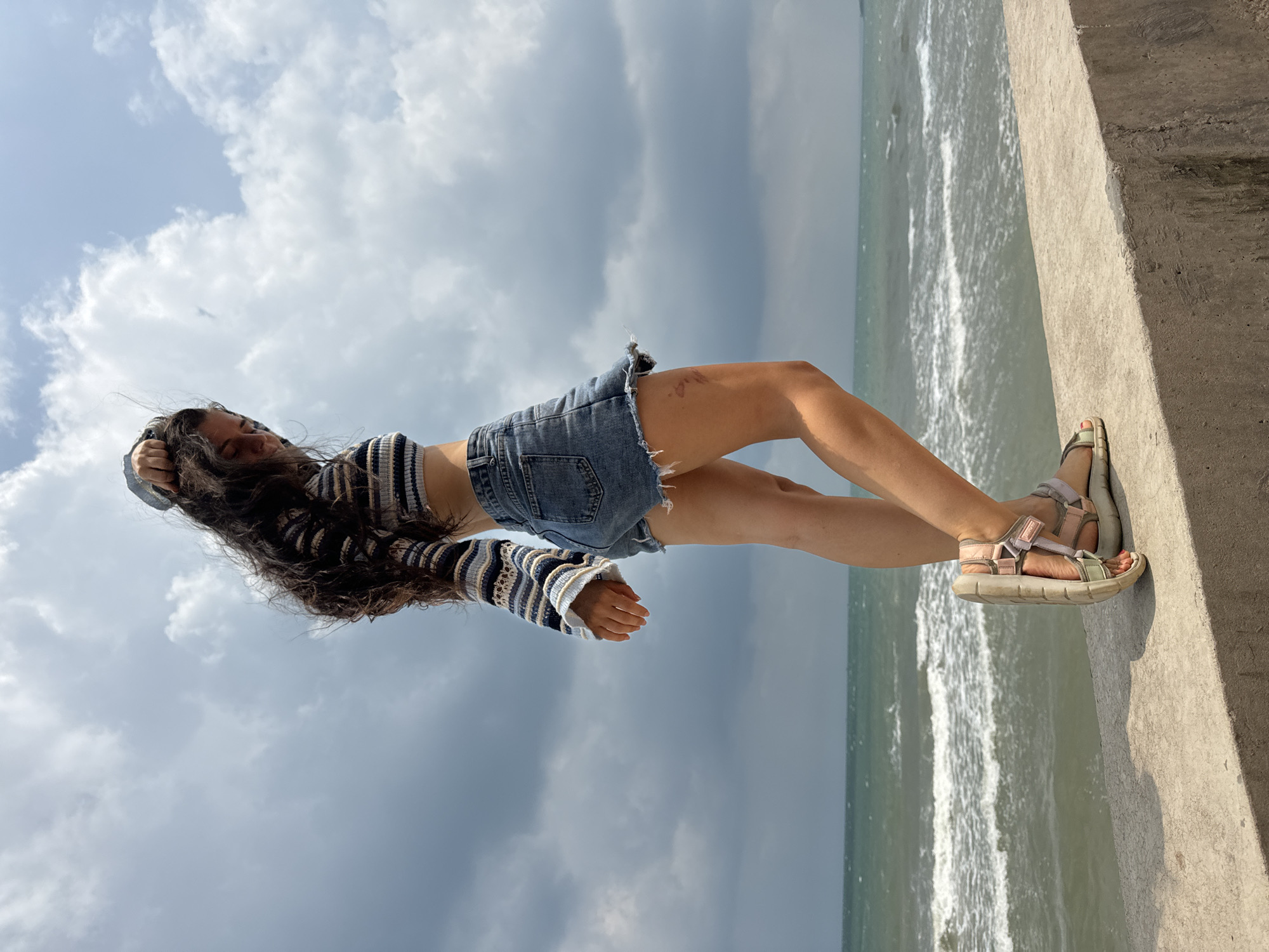 A young woman with long, dark, wavy hair stands on a concrete ledge overlooking the ocean under a cloudy sky. She is wearing a striped crop top, denim shorts, and sandals. Her right hand is raised to her head, and her body is angled away from the camera. The ocean has white-capped waves, and the sky is filled with dramatic, dark clouds. The location is not specified as Phu Thuy Ward, Vietnam.