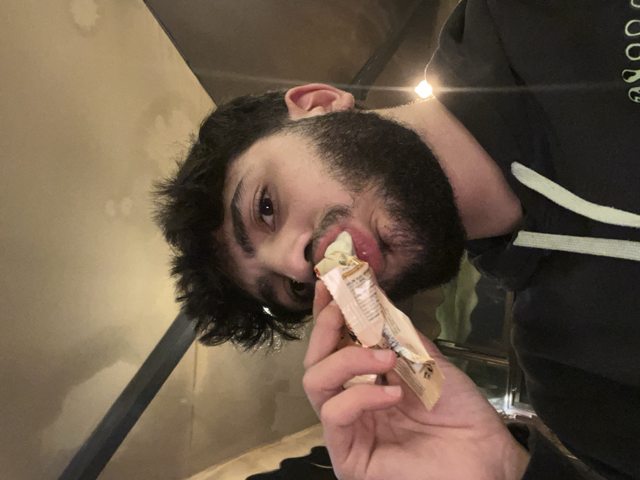 A man with dark hair and a full beard is eating from a beige wrapper. He's holding the wrapper up to his mouth, revealing a bit of the contents. He's wearing a black hoodie with light-colored trim around the hood. The background includes a beige wall with some smudges or stains and a black metal pole. The image appears to be taken from a slightly angled, low-angle perspective.