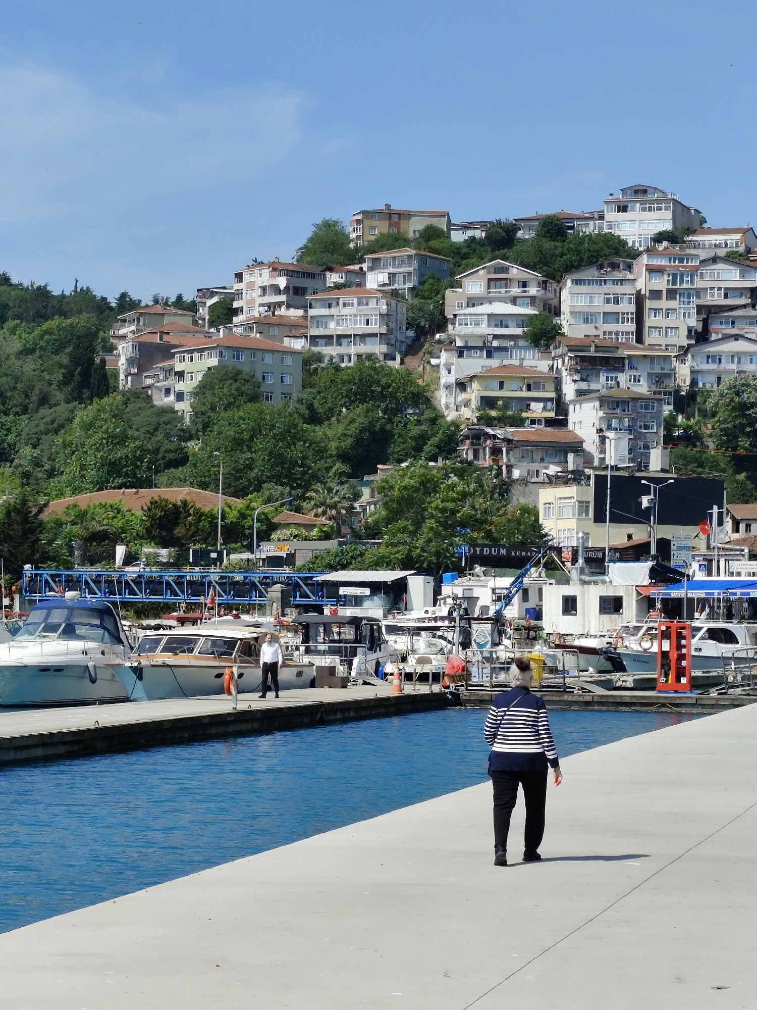 The image depicts a sunny daytime scene in the city of Sarıyer, Turkey. A woman with grey hair, wearing a striped shirt and black pants, walks away from the viewer on a concrete dock. The dock is lined with various boats, including motorboats and smaller vessels. One man, dressed in a white shirt and black pants, stands on the dock near the boats. Behind the dock, a hill is covered with residential buildings and trees. The sky is clear and blue.