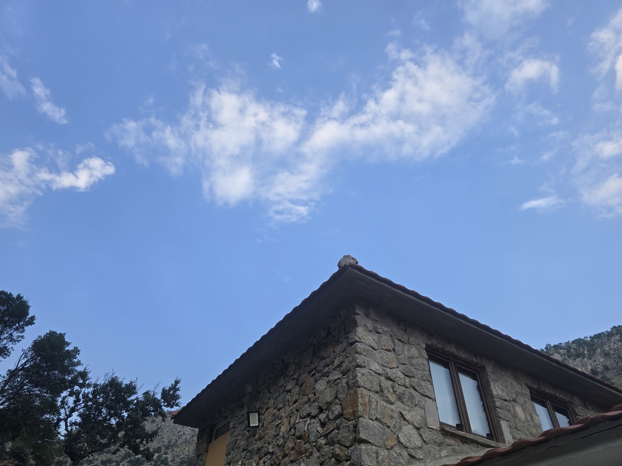 A low-angle shot captures the corner of a stone building under a clear blue sky with scattered white clouds. The building's roof is partially visible, made of dark tiles. The stone walls are constructed from various sizes of rough-hewn rocks. Two rectangular windows with dark frames are set into the wall. A light fixture is mounted on the exterior wall to the left of the windows. To the left of the building, the dark green foliage of trees partially obscures the view. In the background, a rocky hillside with sparse vegetation rises steeply. This scene is likely located in Podgorica, Montenegro, given the typical architecture and natural landscape often found in the region.