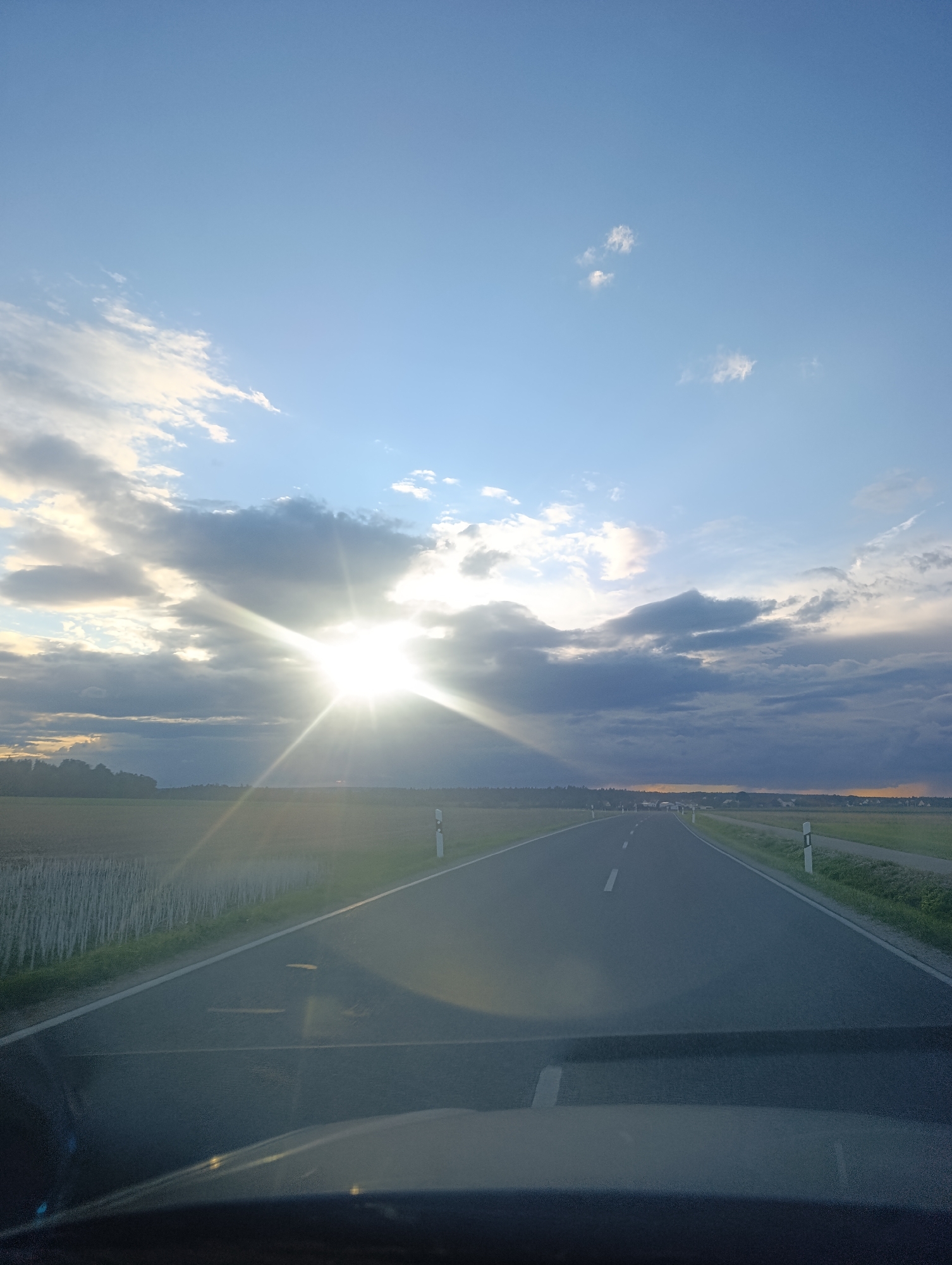 This is a photograph taken from inside a vehicle, looking out over a road in Berching, Germany. The sun is shining brightly through dramatic clouds, casting a lens flare and creating a radiant effect. The road ahead appears to be a two-lane highway, with white lines marking the lanes. Fields of dry stalks line the left side of the road, with a line of trees in the distance. On the right side of the road, there are grassy fields and white road markers. The sky is a mix of blue and grey clouds, with the sun a brilliant focal point. The overall impression is one of a scenic drive through the German countryside during a somewhat overcast but sunny day.