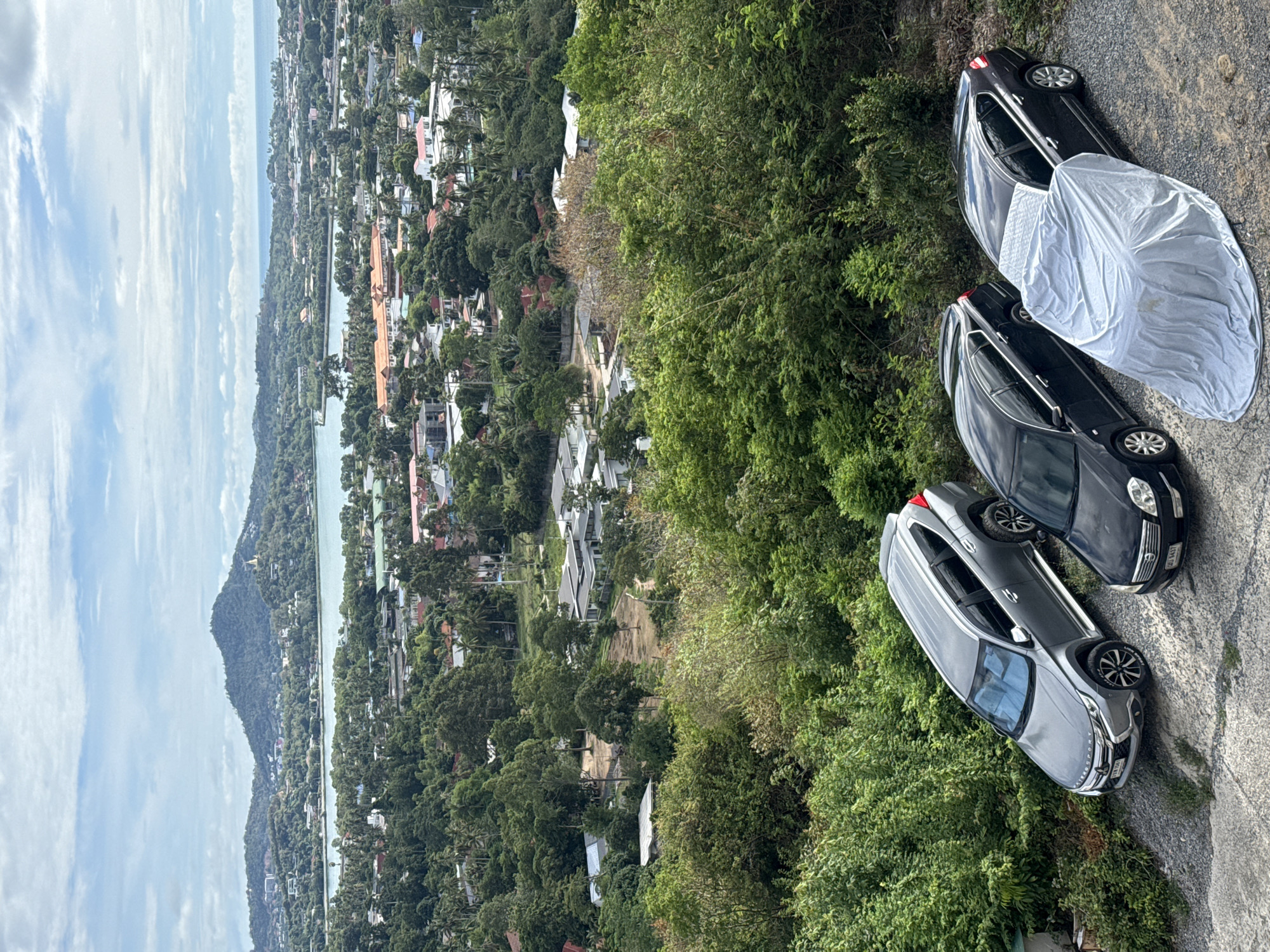 The image depicts a panoramic view from a hillside overlooking a coastal town in Ko Samui District, Thailand. In the foreground, three parked vehicles are visible on a gravel surface. The leftmost vehicle is a gray SUV, followed by a black sedan, and another black sedan covered with a gray tarp. Behind the vehicles, dense green foliage covers the hillside. Further in the distance, a town with buildings and a body of water are spread out, leading to the ocean and a hazy horizon under a cloudy sky. The overall scene conveys a serene, elevated perspective of a tropical, developed coastal area.