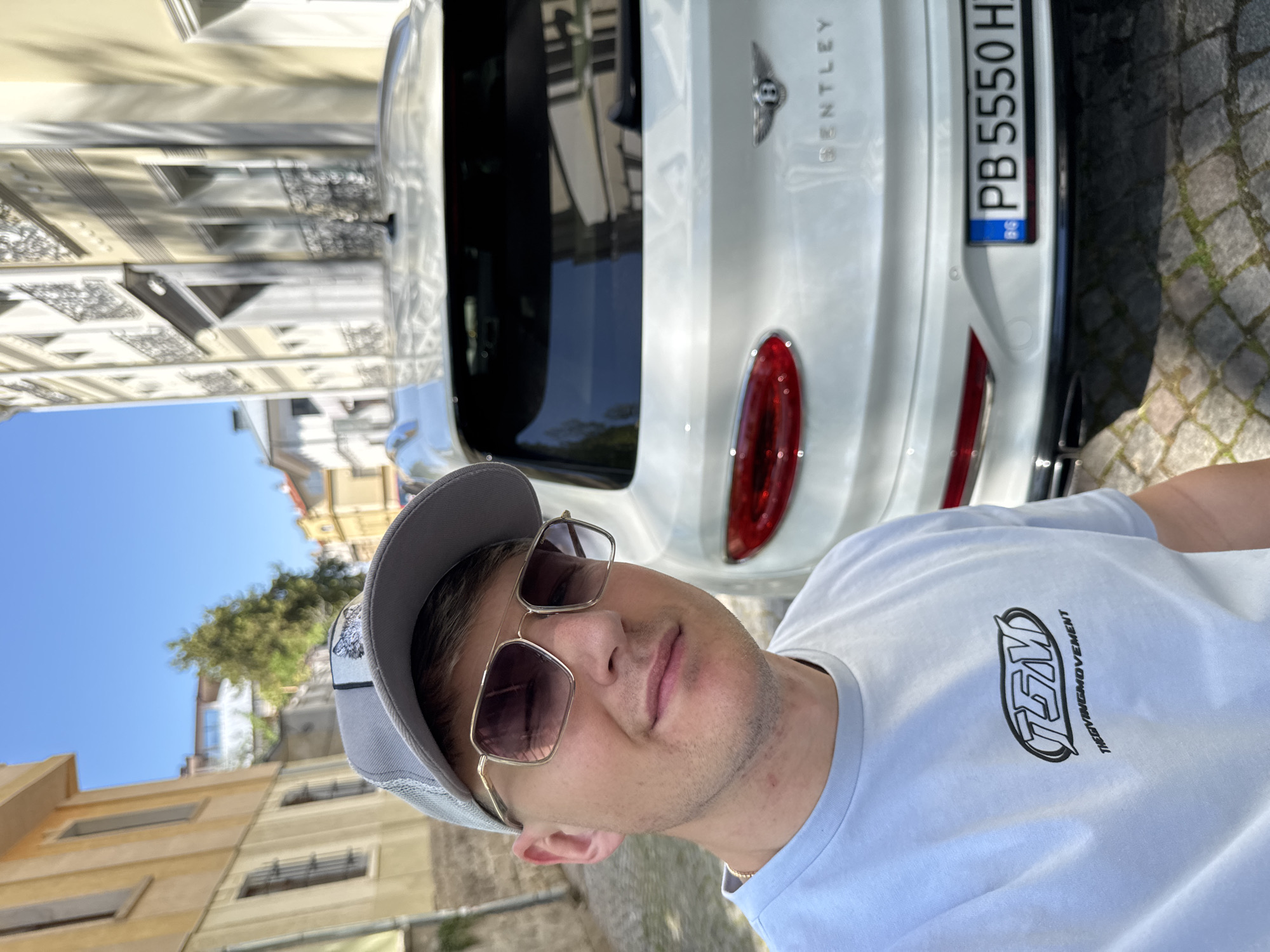 A selfie portrait of a man in a gray cap, gold-framed sunglasses, and a white t-shirt with the "TSM" logo. Behind him is the rear of a white Bentley car with the license plate "PB 5550 H" and a Bulgarian flag on the plate. The location is in Plovdiv, Bulgaria, as evidenced by the Bulgarian license plate and the architectural style of the buildings in the background. The surrounding environment shows buildings typical of a European city, with light-colored facades and wrought-iron balconies. The weather is sunny, casting shadows in the scene.