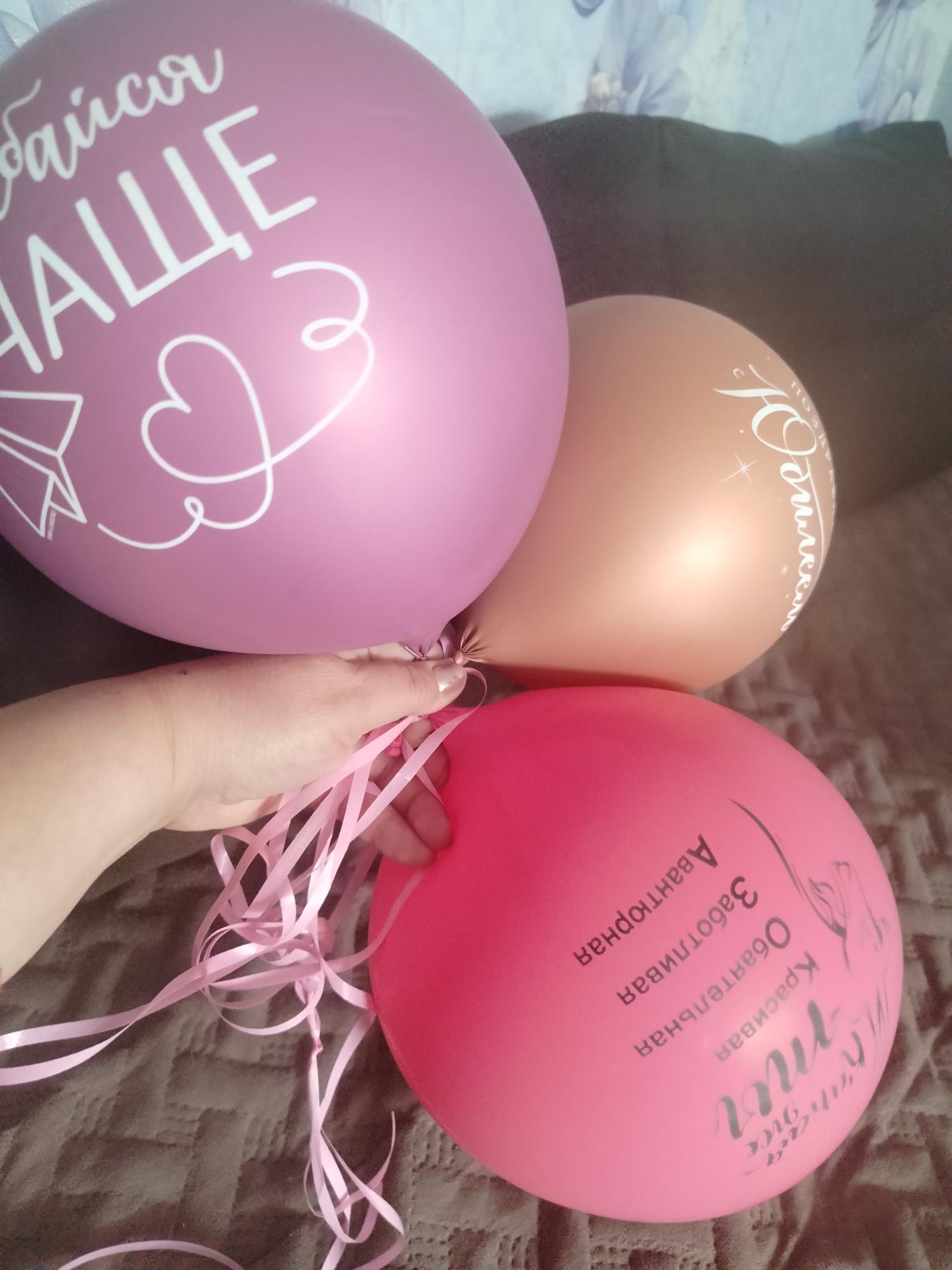 A hand holds a bouquet of three balloons, two pink and one rose gold. The balloons are decorated with white lettering. The largest pink balloon at the top left has the Cyrillic text "байся НАЩЕ" which translates to "be afraid of OUR". There is also a drawing of a heart and a paper airplane on this balloon. The rose gold balloon in the middle has the Cyrillic text "юбилея" which translates to "anniversary". The bottom pink balloon has Cyrillic text that appears to say "Красивая Заботливая Авантюрная" translating to "Beautiful Caring Adventurous" and "my miss". The balloons are resting on a brown textured surface, possibly a bed or sofa. The background is blurred and shows a wall with a floral pattern. The image appears to be taken indoors. There is no identifiable location in the city of Yashkino, Russia.