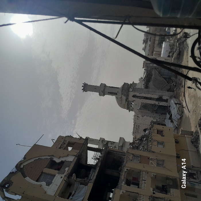 Extensive destruction is visible in an urban setting. Buildings are heavily damaged, some reduced to rubble. A mosque stands amidst the devastation, its minaret still upright but bearing visible signs of conflict. The sky is overcast, adding to the somber atmosphere. The scene suggests recent warfare or disaster, with scattered debris throughout the area. The photo was taken with a Samsung Galaxy A14 phone.