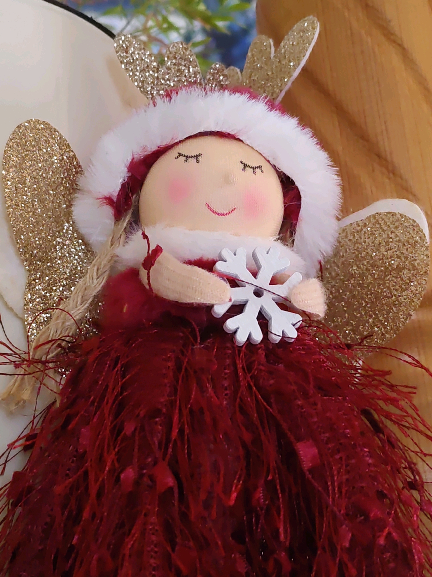 This is a close-up, head-and-shoulders shot of a stuffed holiday ornament, likely a Christmas angel or fairy. The ornament has a round, light-colored fabric face with closed, embroidered eyes and rosy cheeks. It wears a hood trimmed with white faux fur and a pink lining. Attached to the hood are two gold, glitter-covered reindeer antlers. The ornament has gold, glitter-covered wings on either side of its head. It is dressed in a red feathered skirt and holds a white wooden snowflake in its hands. The background is blurred, with a hint of greenery and wood grain. There is no indication of the city of Rostov-on-Don, Russia, or any urban environment.