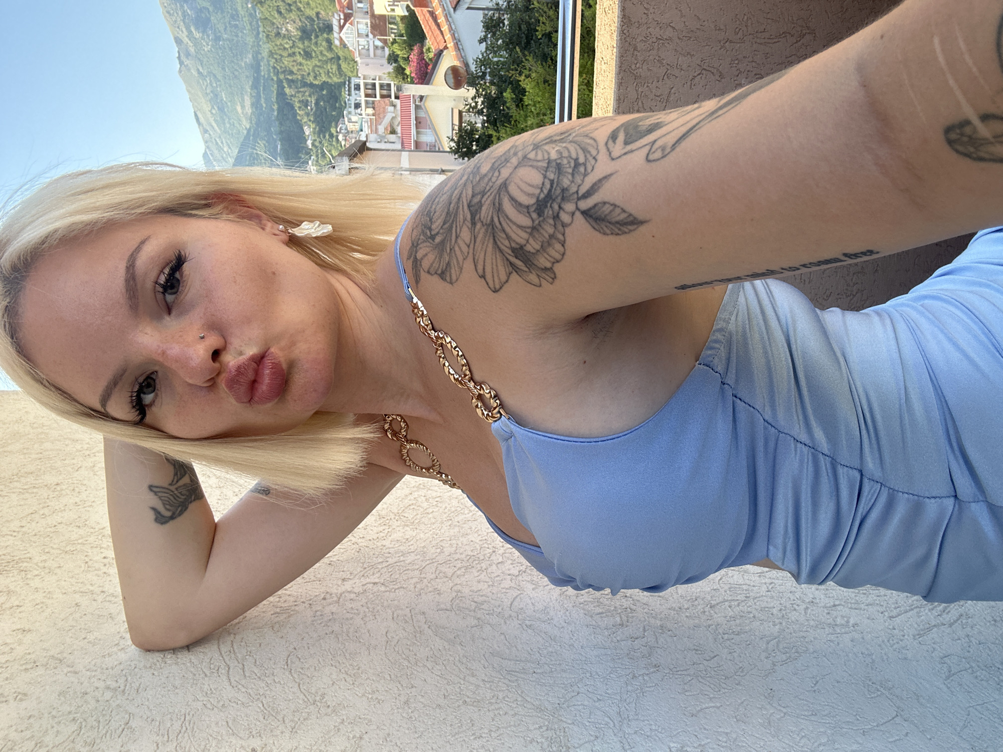 A young woman with blonde hair, wearing a light blue dress with a gold chain detail, poses for a selfie. She has tattoos on her arm and shoulder. Her facial expression is playful, with pursed lips. In the background, a hillside town with red-tiled roofs and lush greenery is visible, set against a clear blue sky. The sun appears to be shining, suggesting it is daytime. The scene is outdoors, likely on a balcony or terrace, with a textured wall in the foreground. The architecture and landscape suggest a Mediterranean or coastal location, consistent with Budva, Montenegro.