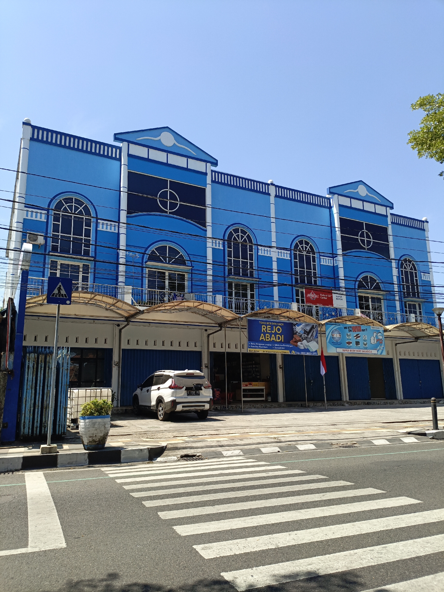 This is a full-shot, eye-level, exterior photograph taken on a bright, clear day of a blue and white commercial building in Salatiga, Indonesia. The building features three stories with arched windows on the upper floors and storefronts on the ground level. Several signs are visible above the storefronts, including one that reads "REJO ABADI" and another that says "WIN". A white SUV is parked in front of the building. In the foreground, a pedestrian crosswalk spans across a paved road. The scene depicts a typical urban commercial street.
