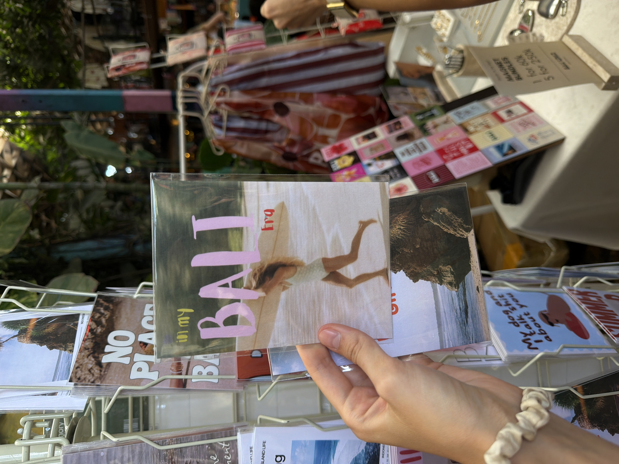 A medium shot captures a person's hand holding a postcard with the text "Bali" in large pink letters, set against a backdrop of a beach with a woman on a surfboard. The postcard is one of many displayed on a wire rack. In the background, there is a table with a display of magnets, likely for sale. The setting appears to be a shop or market, possibly in the city of Kuta Selatan, Indonesia, suggested by the postcards.