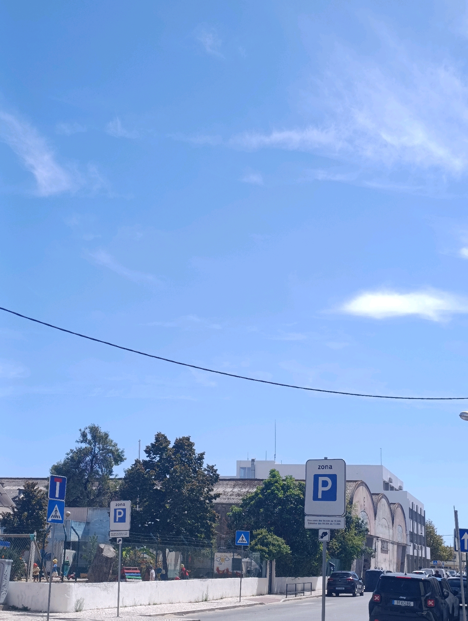 The image depicts a street scene in Setúbal, Portugal. In the foreground, a sidewalk runs alongside a street where several parked cars are visible, including a black Jeep with license plate 91XQ86. A traffic sign indicates a parking zone, labeled "zona P," with operational hours detailed below. Further down the street, more vehicles and pedestrian crossing signs are present. Behind the sidewalk, a playground area is discernible, featuring playground equipment, fencing, and several colorful figures. Mature trees with dense foliage line the perimeter of the playground, and beyond them, a building with arched windows and a white facade dominates the background. The sky above is a clear blue with scattered white clouds and a power line stretches horizontally across the upper portion of the frame.