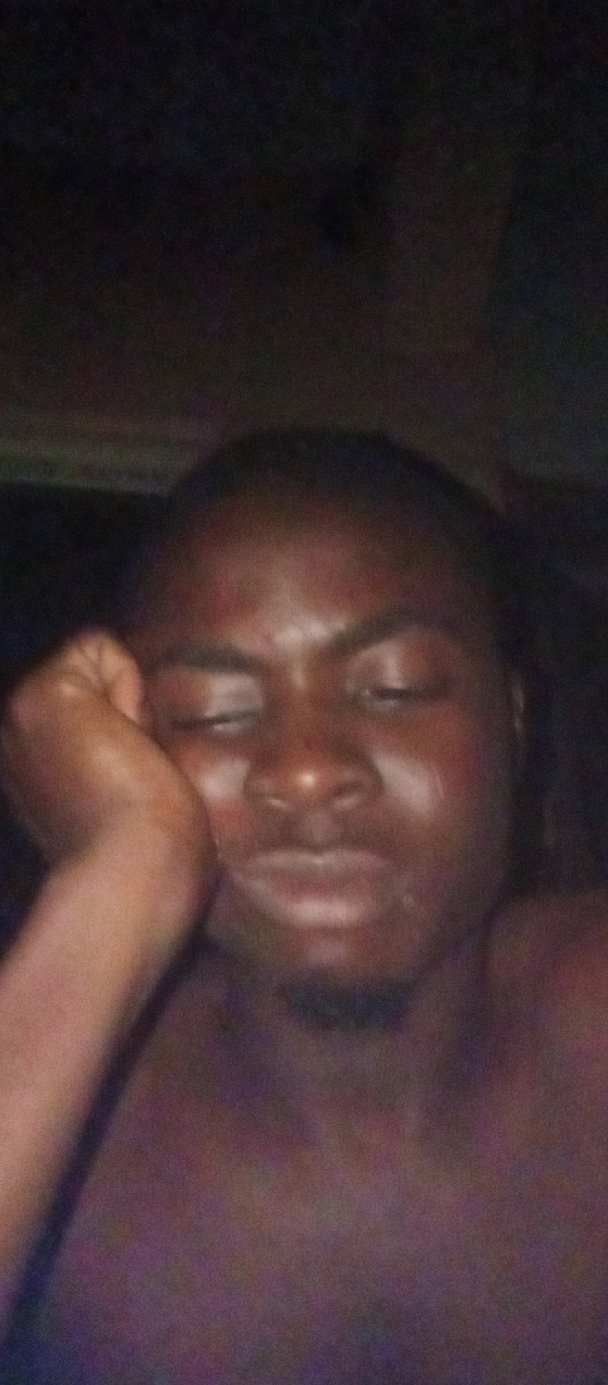 The media file presents a portrait of a person, likely a young man, captured in an extremely dark and dimly lit environment, suggesting nighttime or a poorly lit indoor setting. The individual has dark skin and appears to be bare-chested or wearing a dark top that blends with the background. His left arm is bent, with his hand supporting his head or cheek. His eyes are partially closed or heavily lidded, and his mouth is slightly open or pursed, conveying an ambiguous expression that could suggest tiredness, weariness, or a relaxed state.
The background is uniformly dark, blurry, and indistinct, lacking any specific features or objects. The image quality is low, characterized by significant digital noise and blur, consistent with a photograph taken with a camera (likely a mobile phone) under challenging low-light conditions. There is no discernible text in the image. The context for the image is Ogidi, Nigeria, although no specific visual cues in the photograph directly indicate this location.