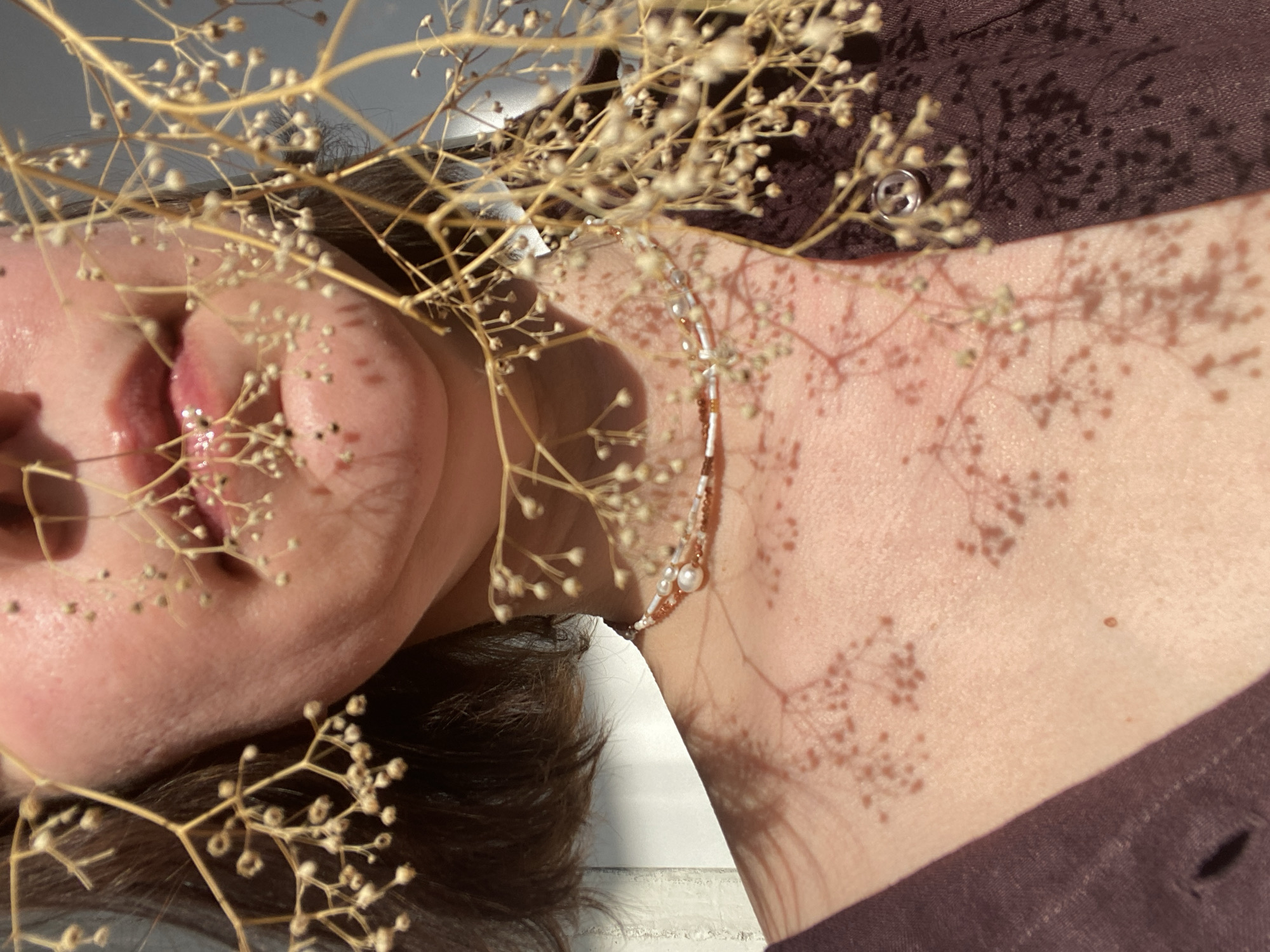 A close-up, artistically composed shot captures a person, likely female, lying down with dried gypsophila (baby's breath) flowers in the foreground and midground. The person's head is oriented to the left, revealing her chin, mouth with slightly parted and glossy lips, and part of her neck. Dark hair is visible near her head and beneath her chin. Around her neck, a delicate beaded necklace or choker with small white and brown/copper beads and a larger pearl-like bead is worn.
She is dressed in a dark brown or maroon garment, possibly a shirt or dress, visible on her shoulder and upper body. Strong, direct sunlight casts intricate, detailed shadows of the dried flowers across her skin and clothing, creating a striking visual pattern. The background is a light-colored, possibly white or grey, surface, suggesting an indoor setting. The sharp shadows indicate a sunny daytime environment, likely during morning or late afternoon. The image conveys a calm, serene, and artistic mood, focusing on texture, light, and natural elements. No text is visible. This image was taken in Chaikovsky, Russia.