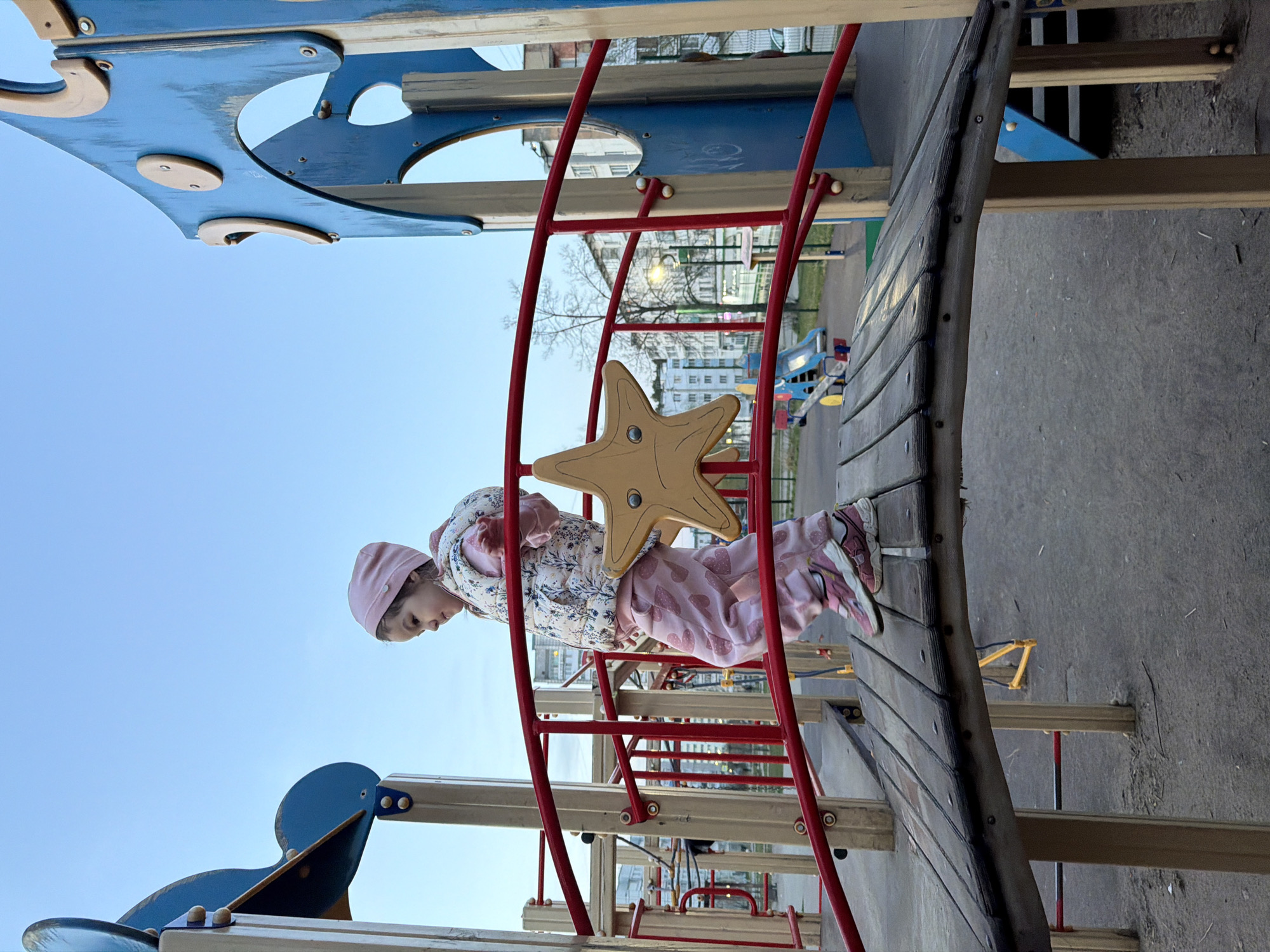 A young child wearing a pink hat, floral jacket, and pink pants climbs on a playground structure in what appears to be the city of Chaikovsky, Russia. The playground has a red ladder-like climbing structure with a yellow star-shaped piece in the middle. The child is partially obscured by the star. There is a blue structure with holes and round elements in the background, as well as a wooden walkway. Buildings and trees are visible in the distance. The sky is a clear blue, suggesting it is daytime. The image is rotated counter-clockwise.