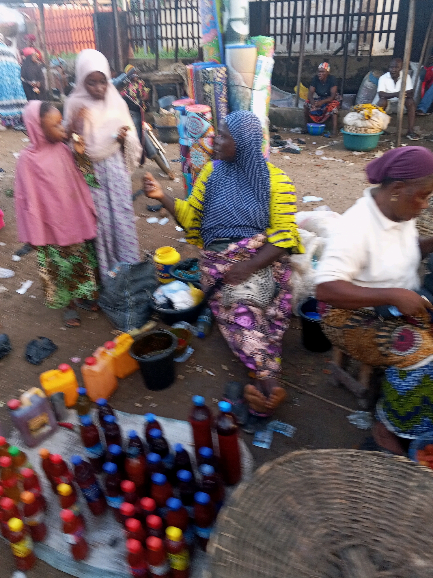 The image depicts a bustling outdoor market scene in Lokoja, Nigeria. In the foreground, a collection of plastic bottles filled with a dark liquid and capped with blue and red are arranged on a light-colored cloth. These bottles appear to be for sale. To the left, two young girls, one wearing a pink headscarf and dress, and another in a lighter pink headscarf and patterned skirt, are present. Behind them, a woman in a light pink headscarf and a patterned skirt is also visible.
Further into the scene, a woman wearing a blue and yellow striped top and a colorful patterned skirt is seated, holding an object in her hand, possibly interacting with the items around her. Adjacent to her, another woman in a white shirt and a patterned skirt is also seated. Several plastic containers, some yellow and orange, are scattered on the ground, along with black plastic bags and buckets. In the background, there are various items, including what appear to be rolls of fabric and other market goods, as well as a metal fence. The ground of the market appears to be dirt. The overall impression is one of a typical marketplace environment with vendors and shoppers engaged in commerce.