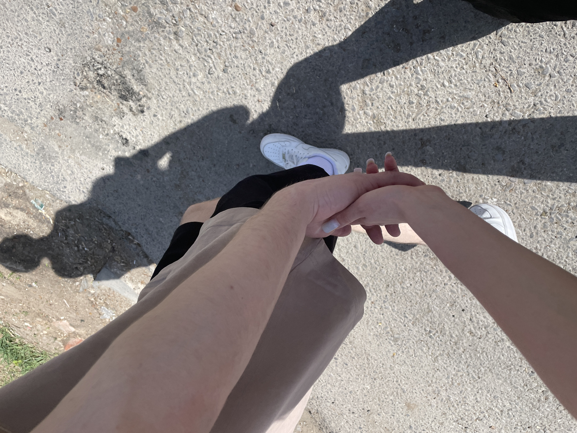 The image depicts a pair of hands clasped together, with one hand belonging to a person wearing a beige garment and the other likely belonging to a person wearing shorts and white trainers, standing on a concrete surface. Shadows cast on the ground suggest an outdoor setting in broad daylight. The location is Ufa, Russia.