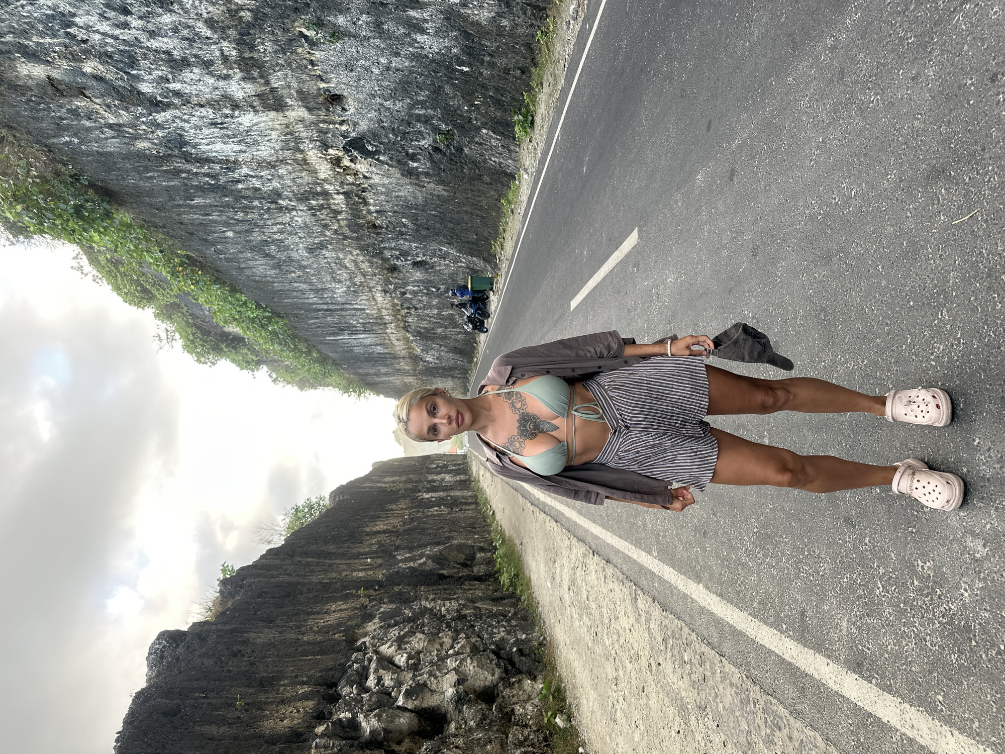 A woman with blonde hair and tattoos on her chest stands on an asphalt road. She is wearing a green bikini top, a grey open shirt, and striped shorts. She is also wearing pink Croc-style shoes and holding a dark hat in her right hand. The road is flanked by tall, rocky cliffs. The sky is overcast. In the background, two people on motorcycles are visible next to a green trash can. The scene appears to be in Kuta Selatan, Indonesia, likely a scenic road with dramatic rock formations.