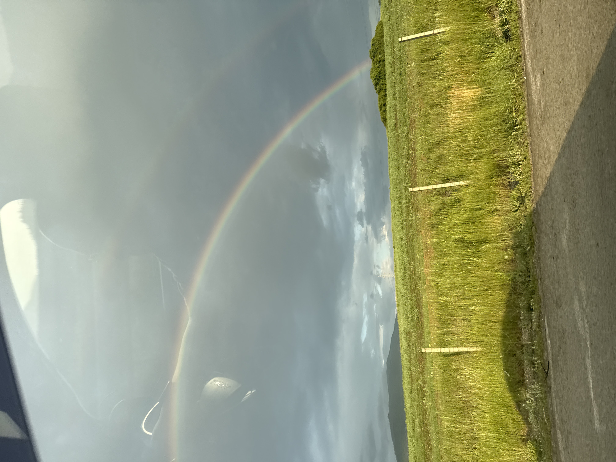 This image captures a rural landscape, likely near Dragichevo, Bulgaria, under a dramatic sky. The scene is dominated by a vibrant primary rainbow arcing from the upper left, descending towards a dense mound of green foliage on a distant hill to the right. A fainter, secondary rainbow is subtly visible above the primary arc, particularly on the left. The sky is largely covered by heavy, dark gray clouds, with patches of lighter sky visible closer to the horizon on the left, illuminating a distant dark mountain range. A thin utility pole is barely discernible on the far right horizon. In the midground, a bright green grassy hillside extends across the frame, with several light-colored wooden fence posts interspersed. The foreground consists of a dark asphalt road, partially obscured by a long, dark shadow cast by the vehicle from which the photograph was taken. A distinct, rounded dark shadow, likely from a car's side mirror or part, is cast onto the green grass embankment on the left. Reflections on a vehicle's window, including an interior car door handle or lever, are visible in the upper left quadrant of the image. No people are visible.