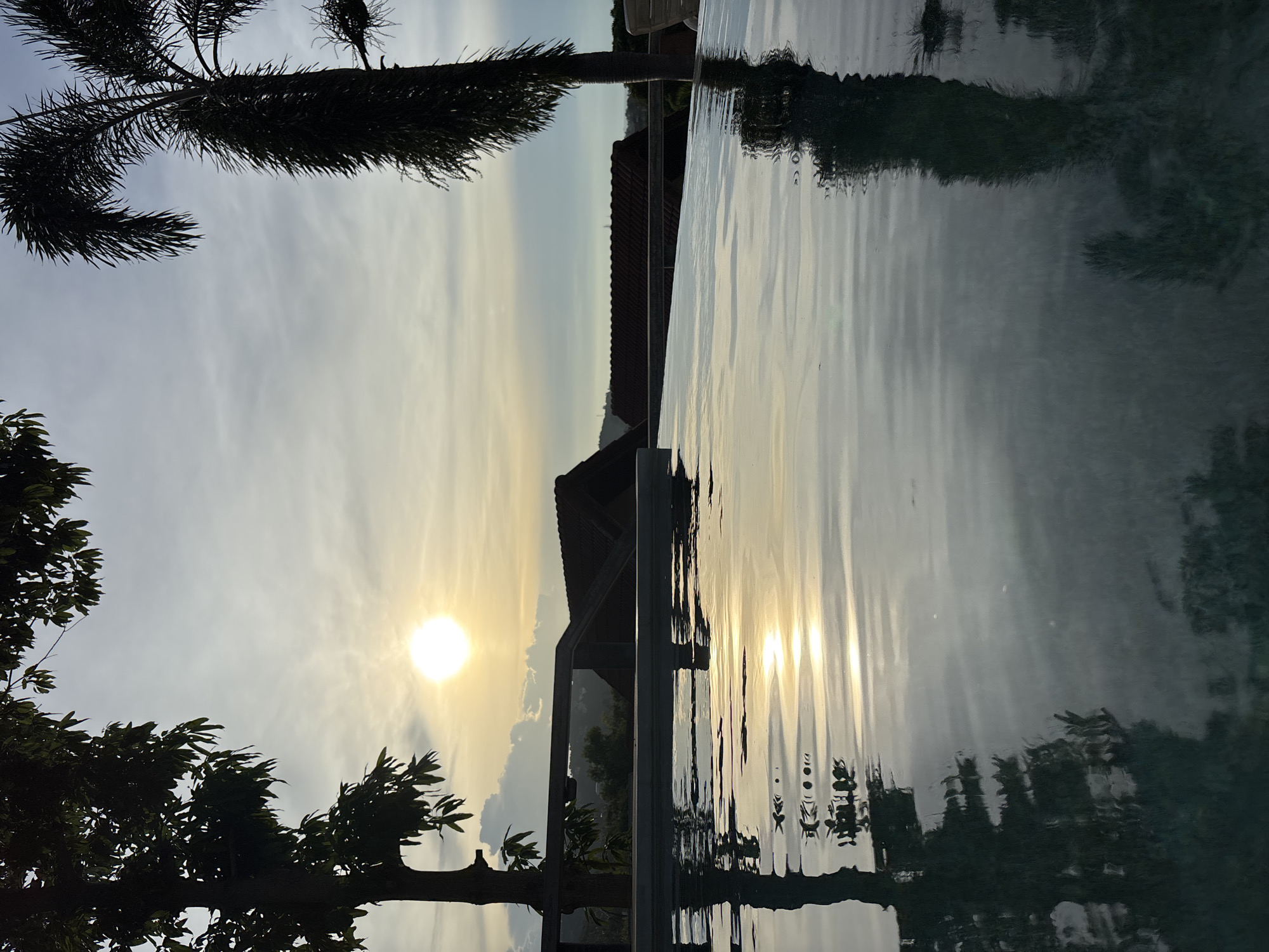 An outdoor scene at dusk shows a swimming pool in the foreground with reflections of trees and the sky. The pool's edge is visible, leading to a blurred background of buildings and distant hills under a hazy, sunlit sky. The sun is low on the horizon, casting a warm glow. A silhouette of a palm tree is on the right, and another tree is on the left. The setting appears to be a resort or residential area in Ban Bang Rak, Thailand.