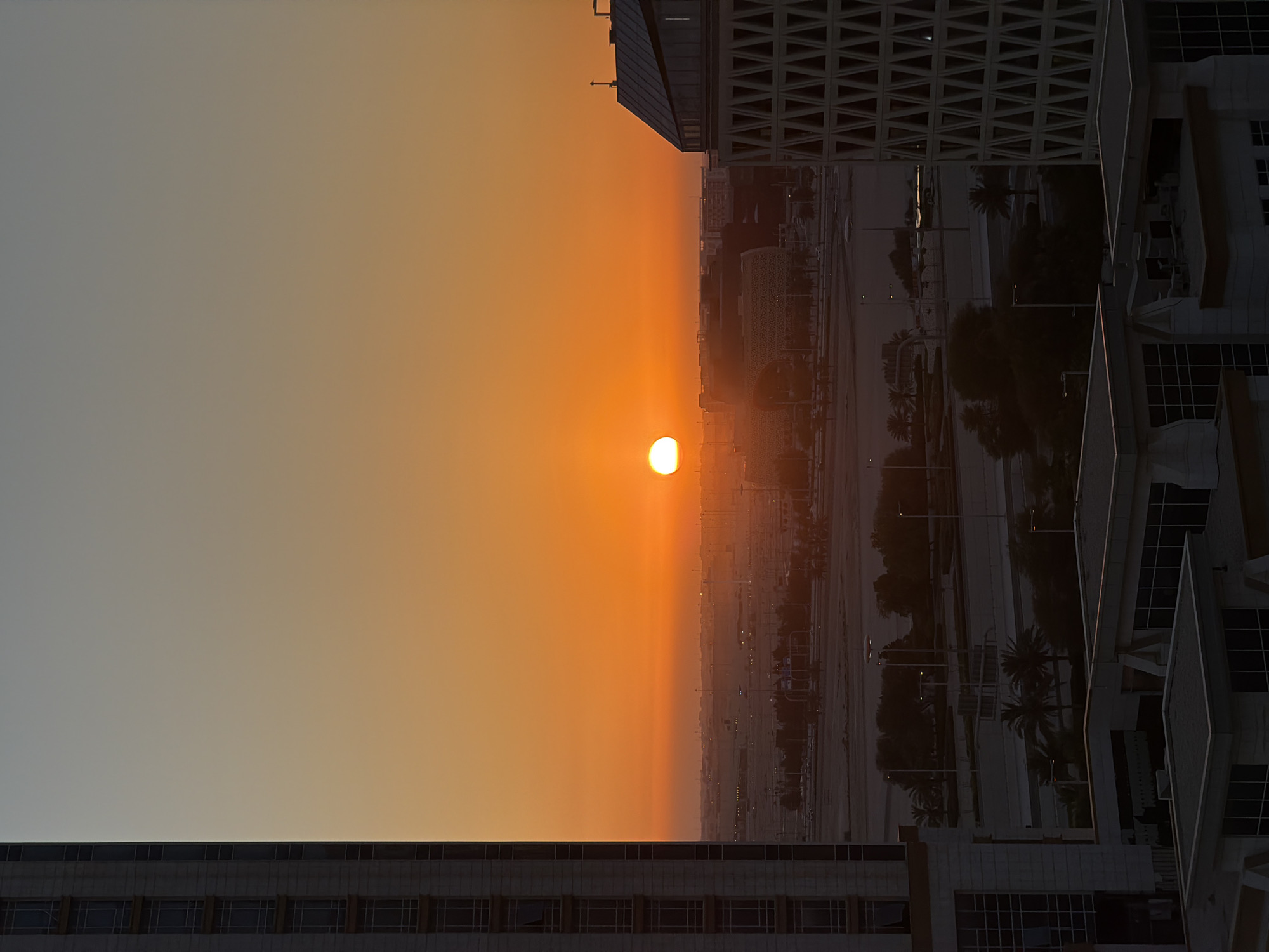 A high-angle, full-shot captures a cityscape at sunset, with the sun as a bright orange orb positioned near the horizon. The sky transitions from a pale gray at the top to a vibrant orange, deepening to a warm yellow just above the horizon. The silhouette of distant buildings and palm trees is visible against the illuminated sky. In the foreground, the facades of two modern buildings are prominent. The building on the left is tall and rectangular with a grid of windows. The building on the right is also modern, with a distinctive geometric pattern on its facade. Roads and streetlights are visible in the mid-ground, suggesting a developed urban environment. The overall scene is bathed in the warm, diffused light of the setting sun.