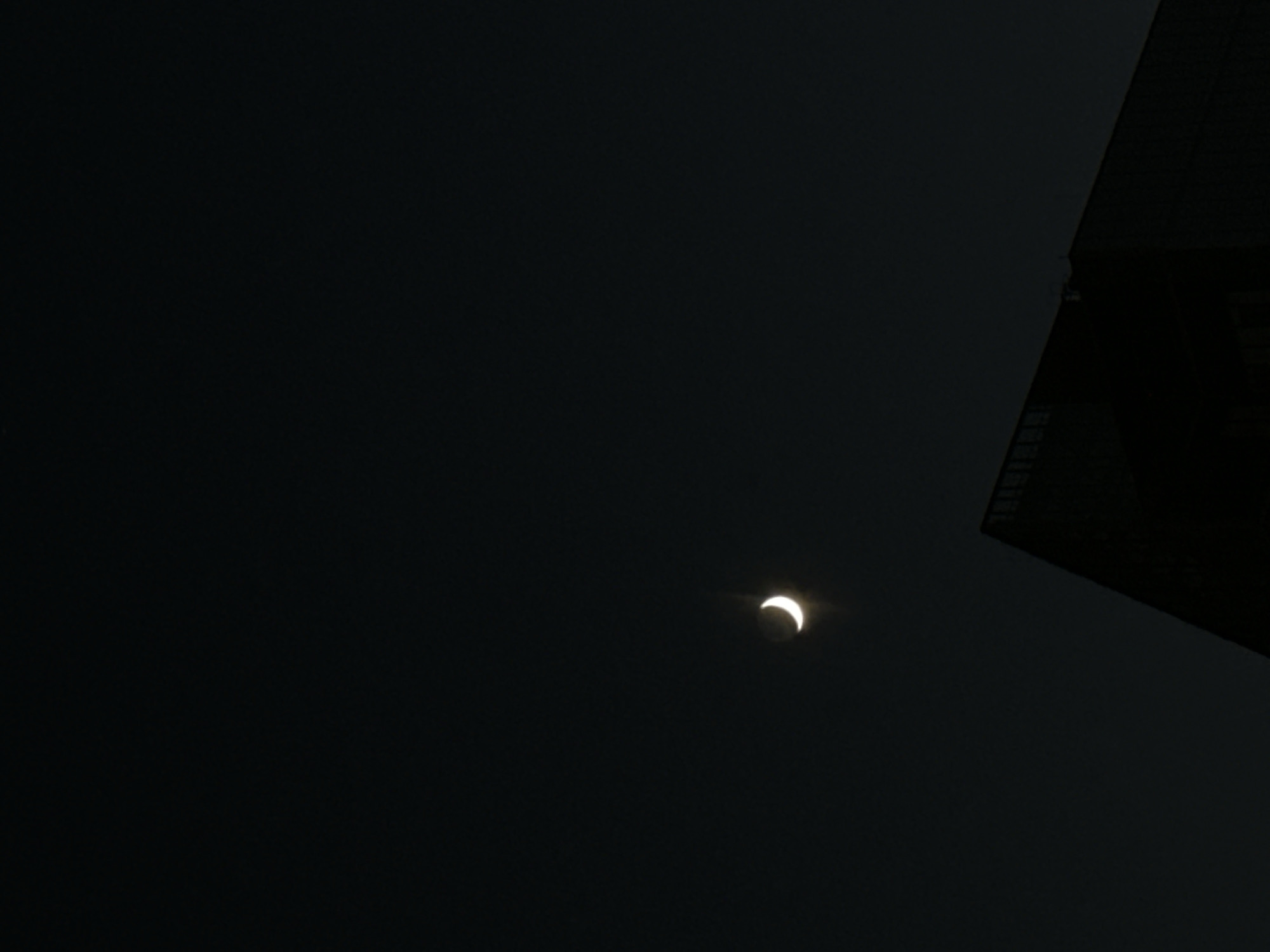 The image is a night time photograph taken in Sofia, Bulgaria. A thin crescent moon is visible in the dark sky, emitting a faint glow. In the foreground, the silhouette of a modern building with a distinctive angled roofline is visible. The building appears to be dark and imposing, with some faint horizontal lines suggesting windows or architectural details on its upper facade. The overall impression is one of urban stillness under a celestial display.
