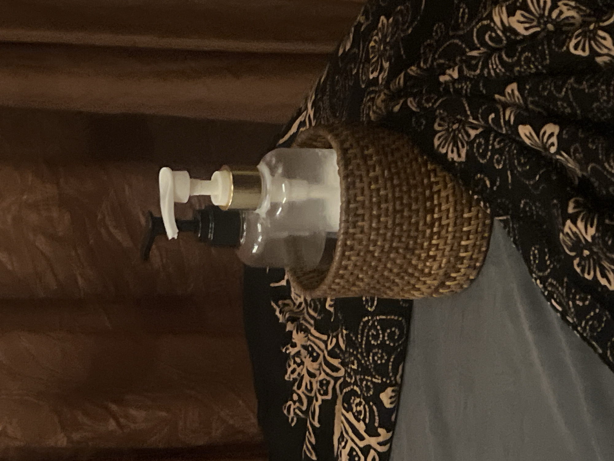 The image, taken in Ubud District, Indonesia, features two pump bottles resting inside a circular, woven wicker holder. The front bottle is clear, equipped with a white pump and a gold-toned collar, while a second, darker bottle with a black pump is partially obscured behind it. These items are set in an indoor environment. On the right side of the frame, a dark fabric, possibly a curtain or bedding, displays an intricate, cream-colored floral and paisley pattern. Below this, a plain grey fabric is visible. To the left, a solid dark brown surface, likely wood paneling or furniture, forms the background. The arrangement suggests a detail from a bathroom or bedroom, possibly within a resort or guesthouse, highlighting local aesthetic elements such as natural woven materials. No individuals are present, and no specific activity is occurring. The lighting appears soft and ambient. No visible text is present on any of the objects.