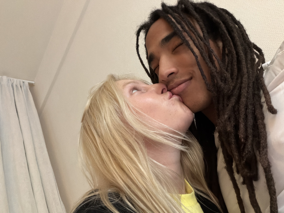 The image displays a close-up, angled shot of two individuals indoors. A fair-skinned individual with long blonde hair, appearing female, is seen on the left, kissing the lips of another individual. Her left eye is open and directed upward. She wears a yellow garment under a dark outer layer. The second individual, on the right, has dark skin and dark dreadlocks, appearing male. His eyes are closed or nearly closed, and his lips show a slight upward turn. Their lips are pressed together in a kiss. The background is a plain, light-colored wall or ceiling. A light-colored, pleated Roman blind or similar window covering is visible in the upper left corner. No specific environmental details are present to identify a precise location within the city of Paris, France.