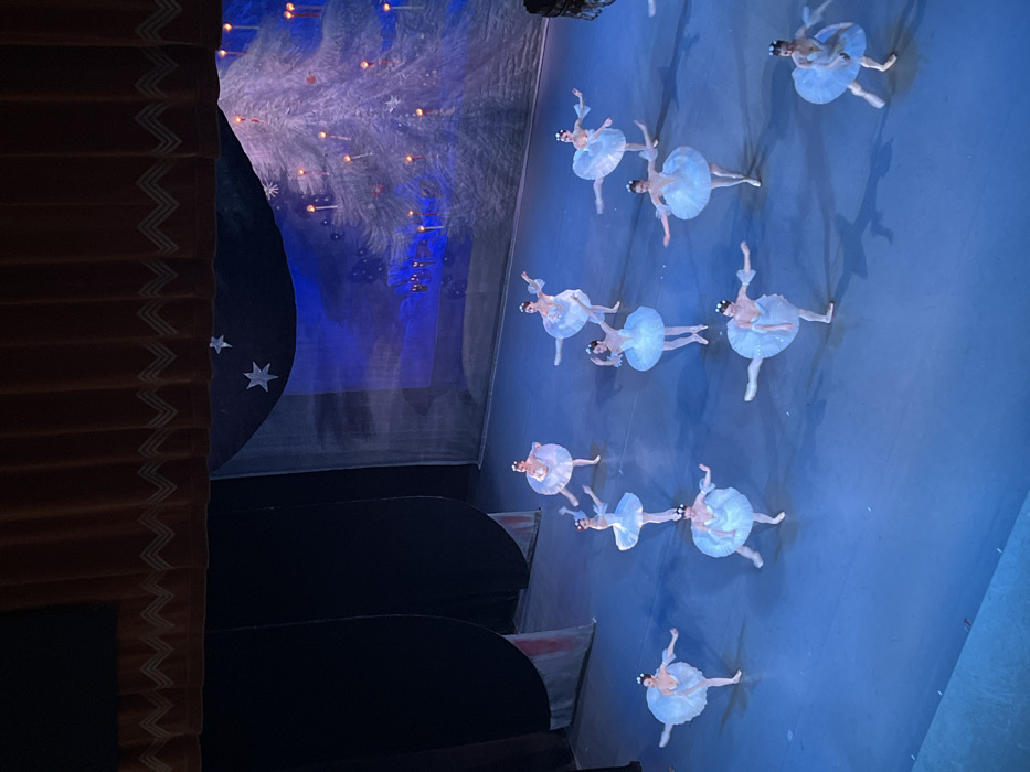 A ballet performance unfolds on stage under blue lighting. Numerous ballerinas in white tutus are arranged in a pattern. The stage backdrop depicts a wintery scene with a decorated tree and sparkling lights. Stage curtains with decorative trim are visible on the left, partially obscuring a set piece decorated with stars.