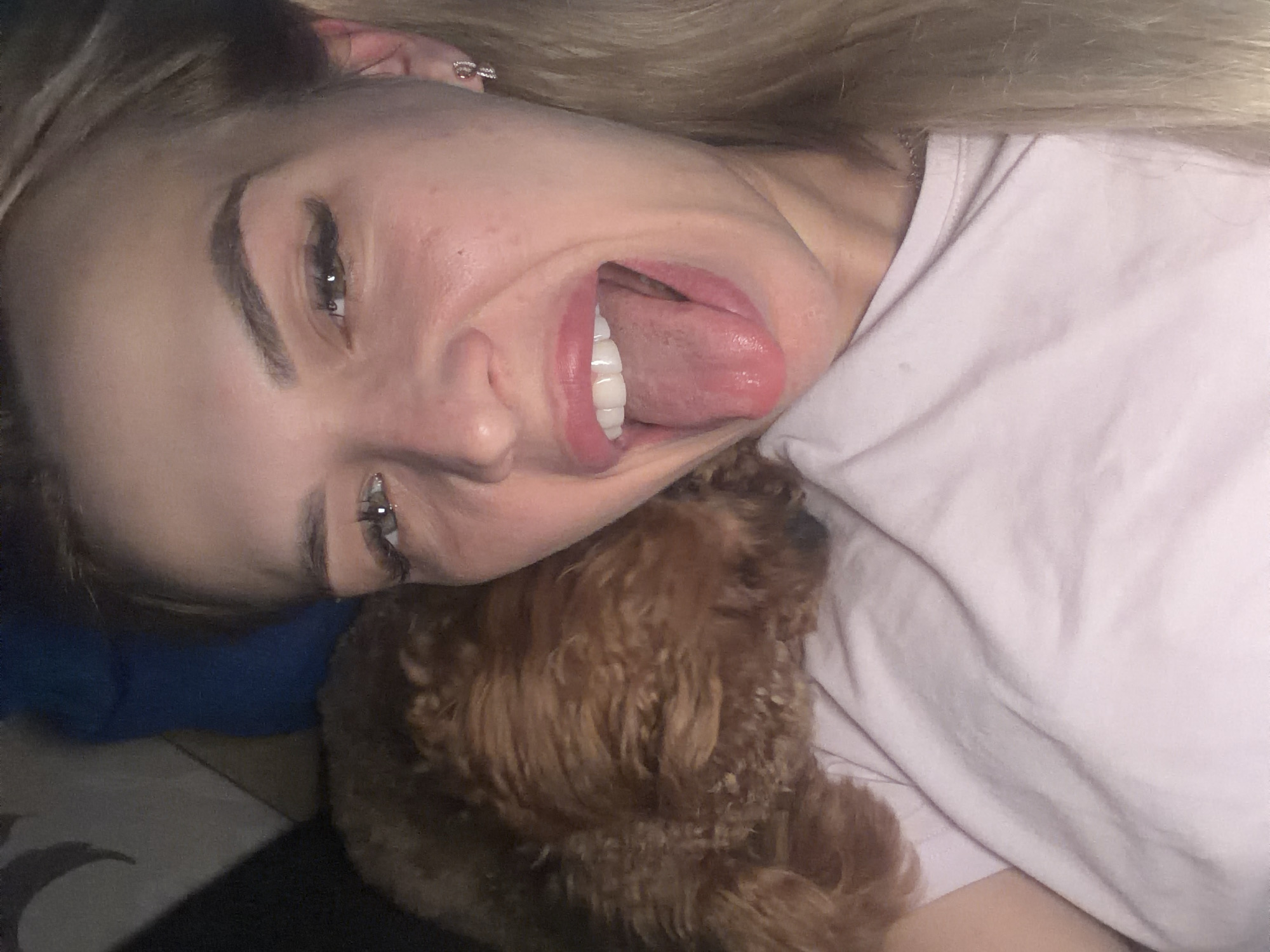 The image is a close-up, selfie-style portrait of a young Caucasian woman with blonde hair and light-colored eyes. She is wearing a light pink t-shirt. Her mouth is open, and her tongue is extended. To her left, partially obscuring her shoulder, is a fluffy, light brown dog, likely a poodle or similar breed. The background appears to be an indoor setting, possibly a bed or couch, with some out-of-focus dark and blue elements. There is no discernible information in the image that would allow for the localization of this scene within the city of Kyiv, Ukraine.