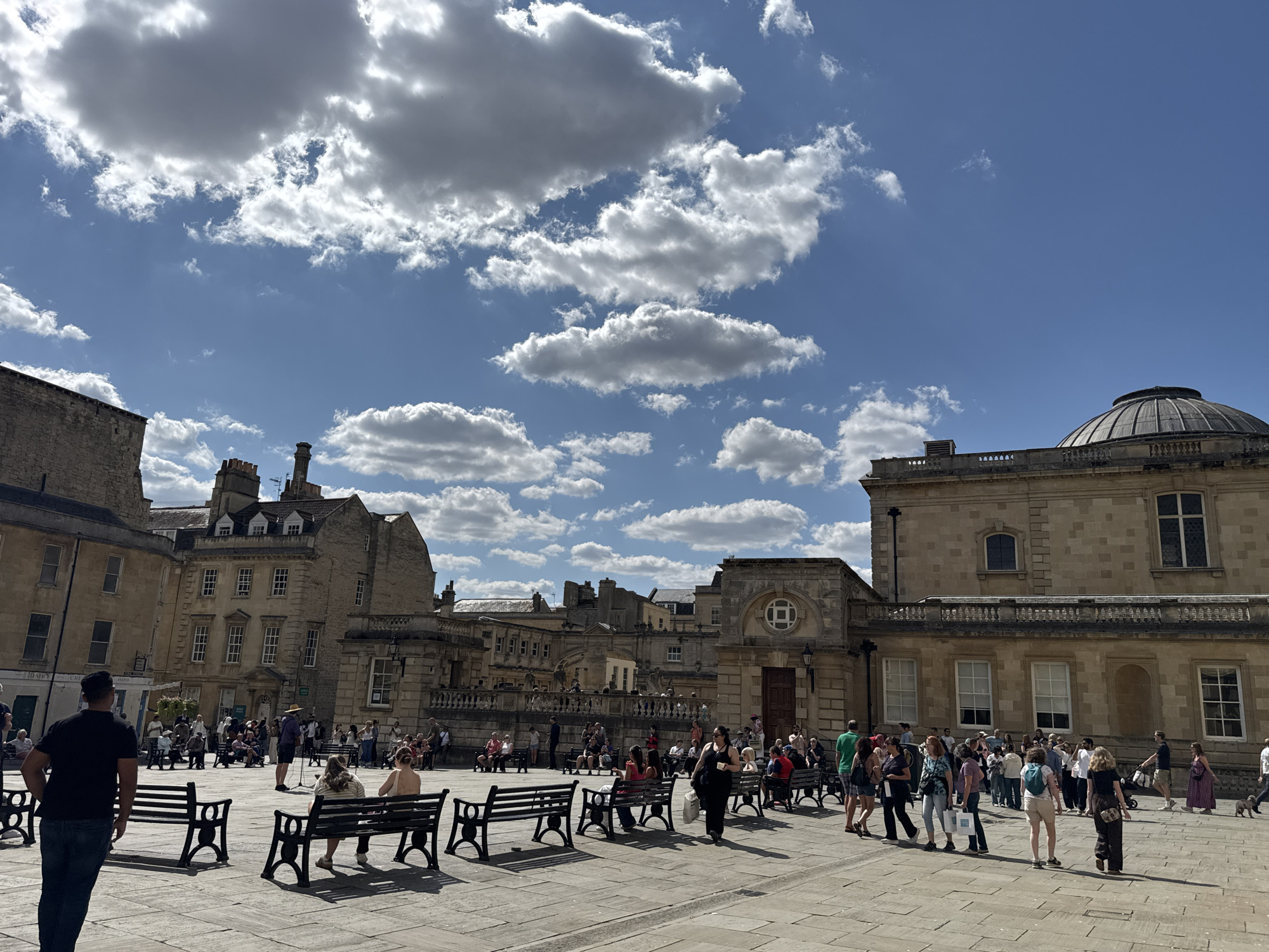 The image captures a view of the Abbey Churchyard in Bath, United Kingdom, on a sunny day with scattered clouds. Several buildings with traditional Georgian architecture surround the open public space, which is paved with stone. A significant portion of the image is occupied by the sky, showcasing a dynamic cloudscape against a bright blue backdrop.
In the foreground, several dark-colored benches are placed on the pavement. Some are occupied by people, while others are empty. A man wearing a black t-shirt and jeans is seen walking away from the camera, towards the left side of the frame.
Further into the scene, a group of people are gathered in the middle of the square, some sitting on benches and others standing. A performer, possibly a street musician, is standing and singing or speaking into a microphone, with a small audience facing him. Many individuals are dressed in casual summer attire.
The background features more of the stone buildings, including one with a prominent dome, characteristic of Bath's architectural style. Balconies and windows are visible on the facades of the buildings. The overall atmosphere is one of a bustling public square on a pleasant day, with tourists and locals enjoying the outdoor space.