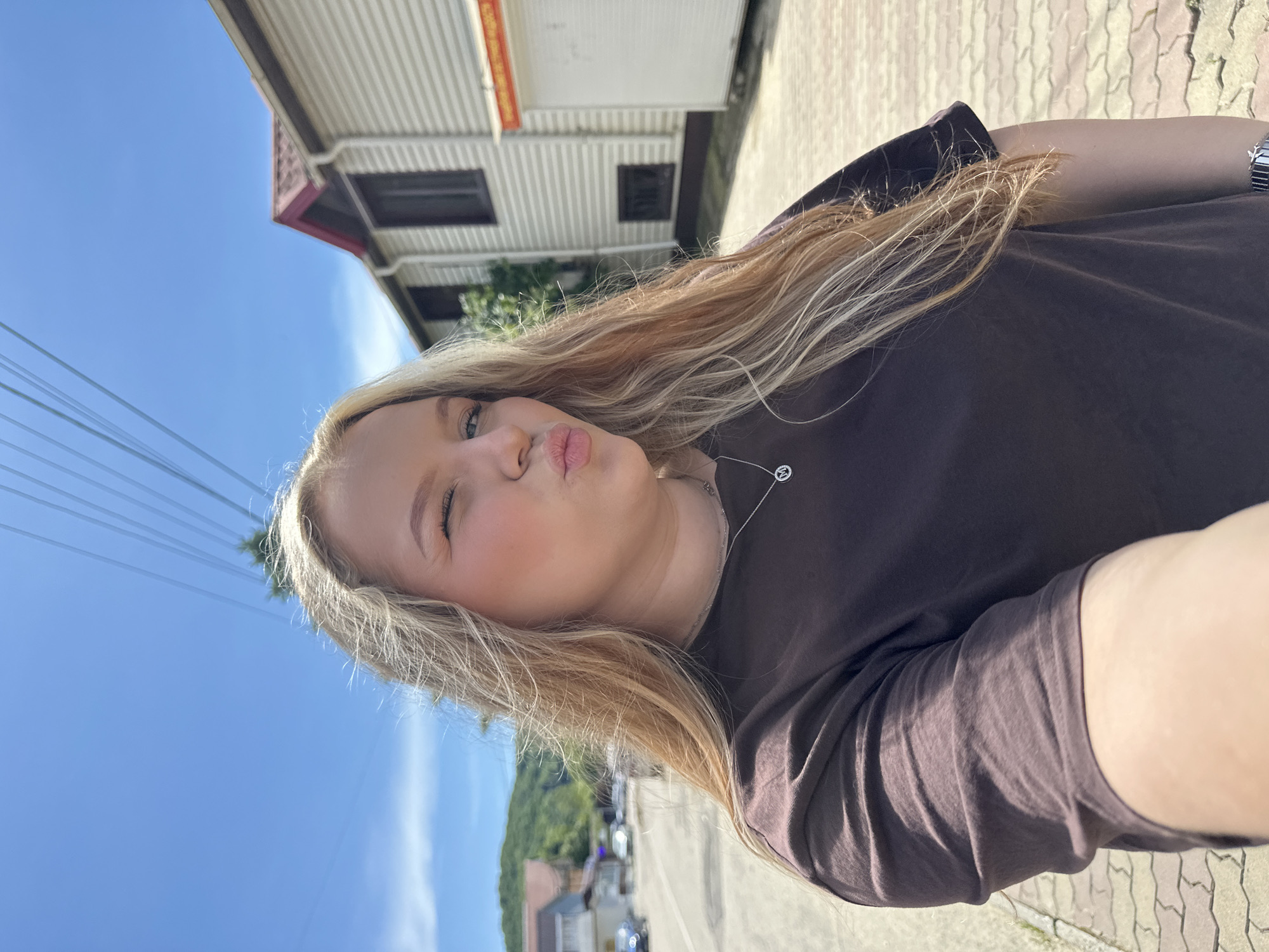 A young woman is featured in a selfie-style portrait, her head and torso dominating the frame, which is significantly tilted. She has long, light blonde hair and fair skin. She is wearing a dark, short-sleeved t-shirt, appearing brown or dark grey, and a delicate silver-colored necklace with a circular pendant that features what resembles a stylized letter 'M' or similar symbol. A metallic watch is visible on her left wrist. Her expression is playful and affectionate, characterized by pursed lips as if blowing a kiss and her right eye winking.
The setting is outdoors under a clear, vibrant blue sky, indicating a bright and sunny day, likely morning or afternoon. To the upper left, several parallel power lines cut across the sky. In the mid-ground, a white two-story building with corrugated siding and a sloped roof with red-orange accents is visible, featuring dark windows. A red sign with illegible text can be seen on its upper facade. Below this building, green foliage and some indistinct parked vehicles are visible along a road or path. The ground is paved with light-colored interlocking bricks. The distant horizon shows forested hills or mountains, suggesting a natural landscape beyond the immediate built environment. The location cues, including the architecture and landscape, are consistent with Lermontovo, Russia. The strong sunlight casts clear shadows, contributing to the bright and cheerful atmosphere.
