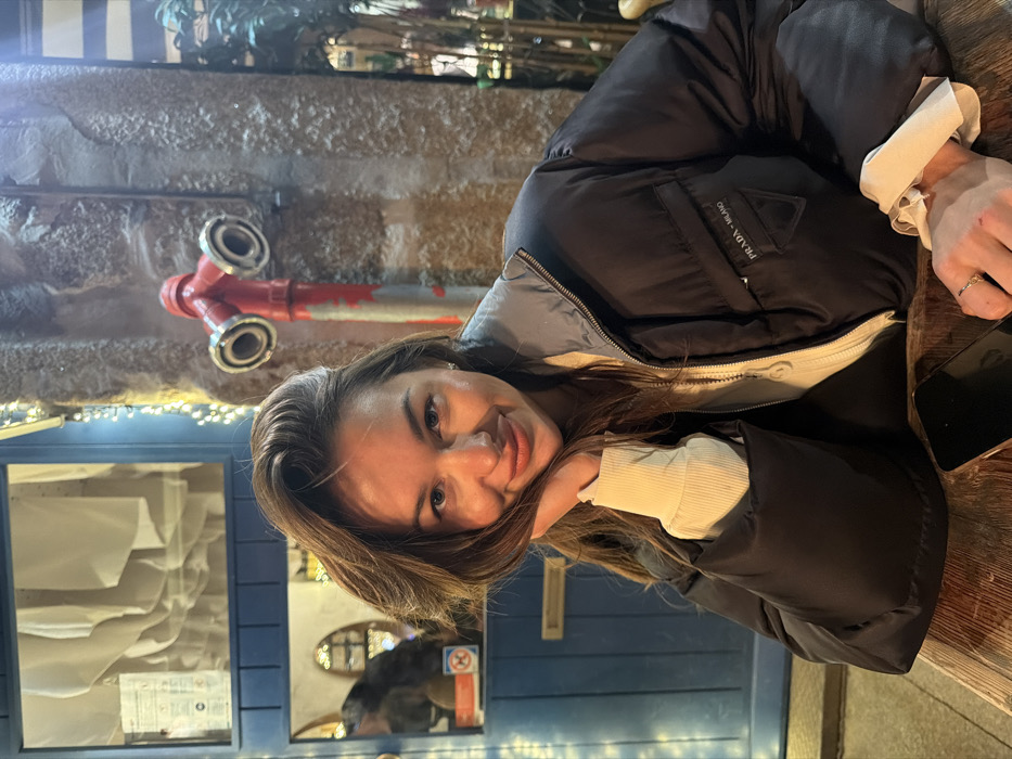 A woman smiles while seated at a wooden table outside. She wears a black puffer jacket with a white ribbed undershirt. The jacket has a Prada logo on the chest. A smartphone lies on the table beside her. Behind her are a blue painted facade with a display window and a stone wall featuring a red fire hydrant. String lights illuminate the background.