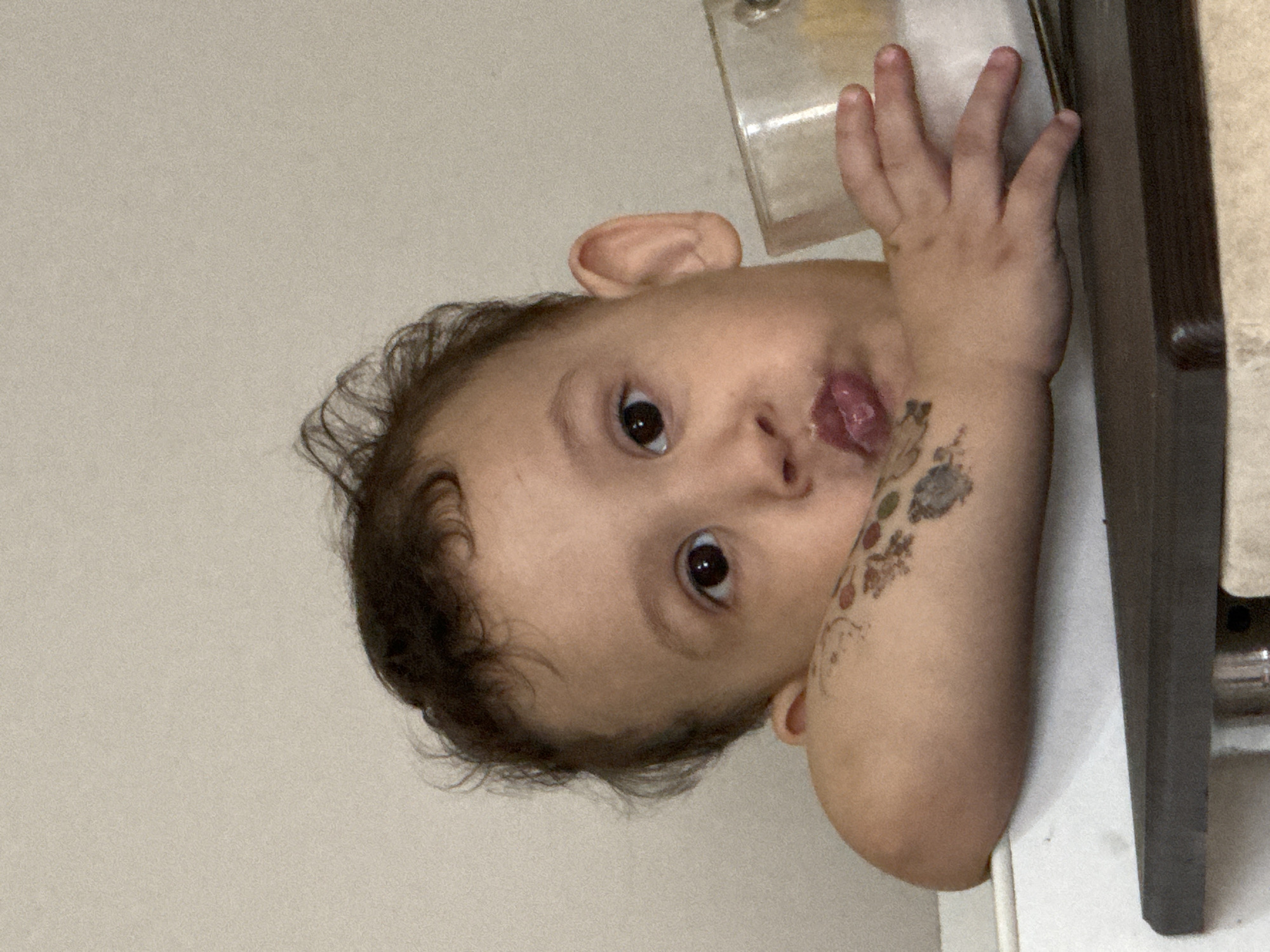 A baby with brown hair is lying on their side. The baby's mouth is open, and their tongue is visible. Their arm is in front of their body, and there is a design on their arm. The baby is next to what appears to be the edge of a counter. The background is a plain, light color. There is no evidence of the city of Chaikovsky, Russia, in the image.