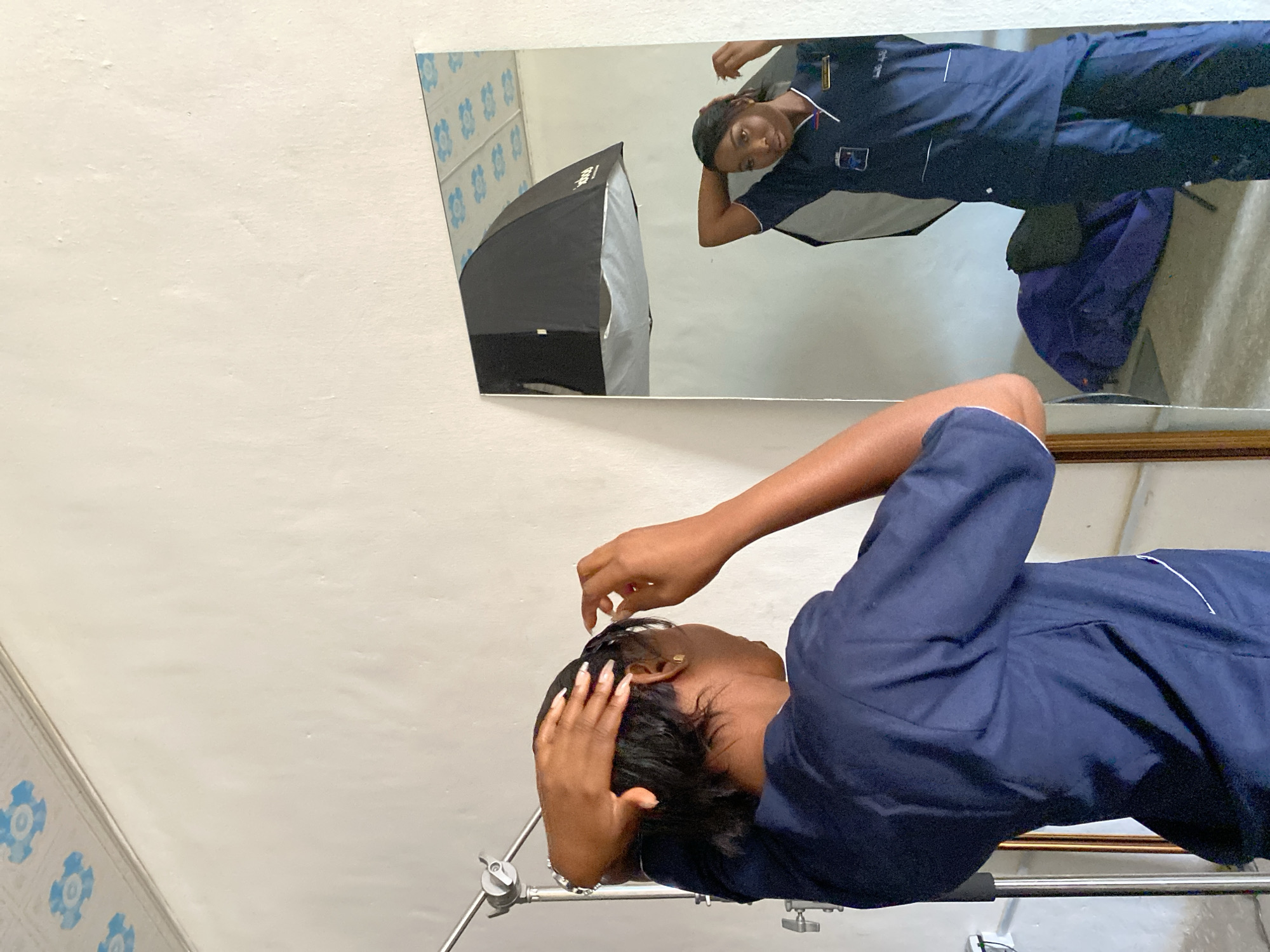 A person in Gura Top, Nigeria is seen in the lower-right of the frame, adjusting their hair. They are wearing a navy blue uniform. Above and to the left of the person is a mirror showing their reflection. The reflection shows the person in full uniform, plus a black softbox. The wall behind the mirror is white with blue decorative floral tiles.