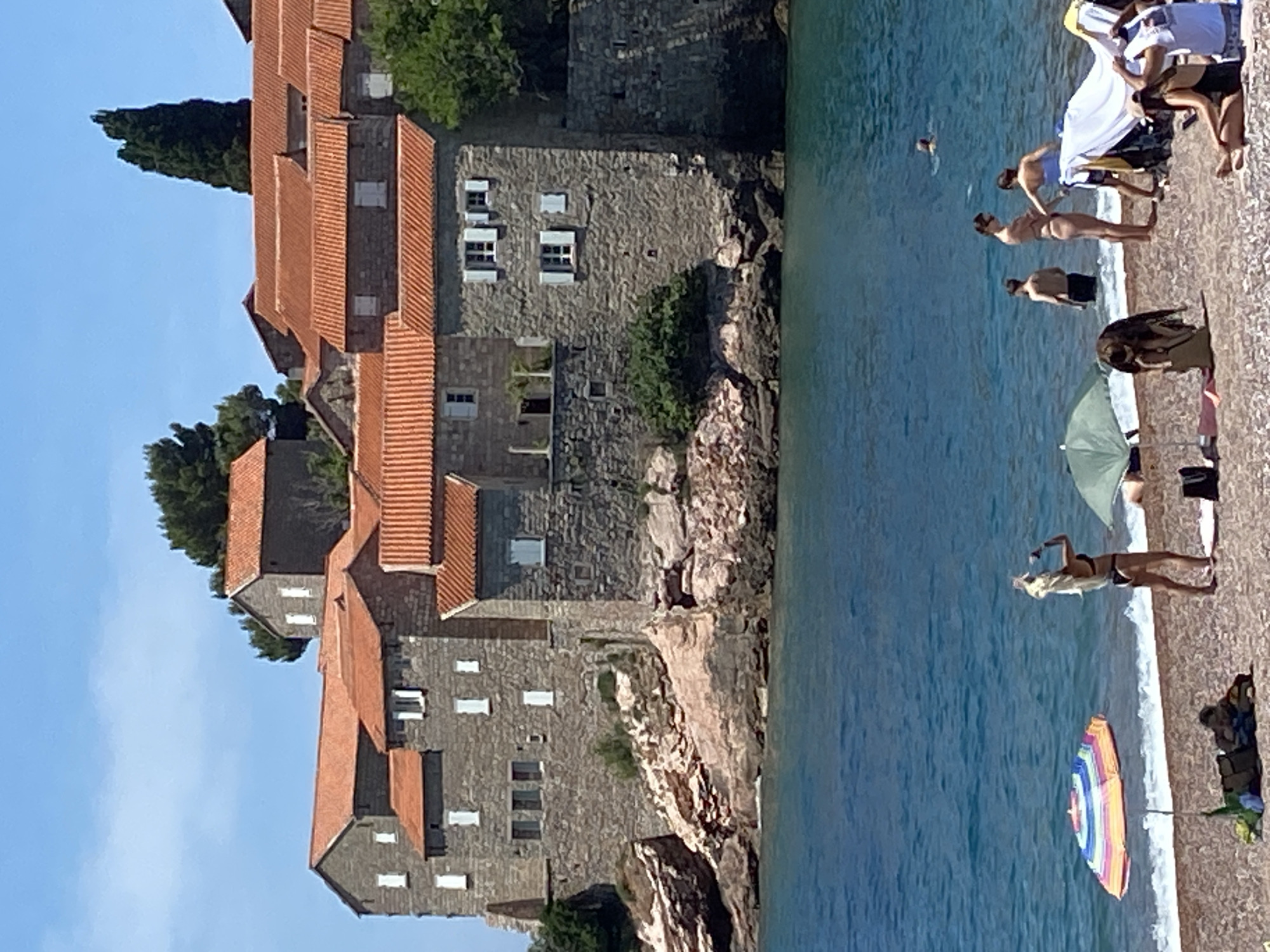 The image depicts a scenic view of Sveti Stefan, Montenegro, with its iconic island resort visible in the background. In the foreground, a beach scene unfolds with several individuals enjoying the water and sand. A woman in a bikini stands near the water's edge, holding what appears to be a smartphone to her ear. Nearby, an open, striped umbrella shades a person lying on a beach towel. Further into the sea, a lone swimmer is visible. The water is a vibrant blue, characteristic of the Adriatic Sea. The island resort itself is a cluster of stone buildings with terracotta roofs, perched on a rocky outcrop. Lush green trees surround the buildings, and the clear blue sky suggests a sunny day. The overall impression is one of a tranquil summer day at a picturesque coastal location.