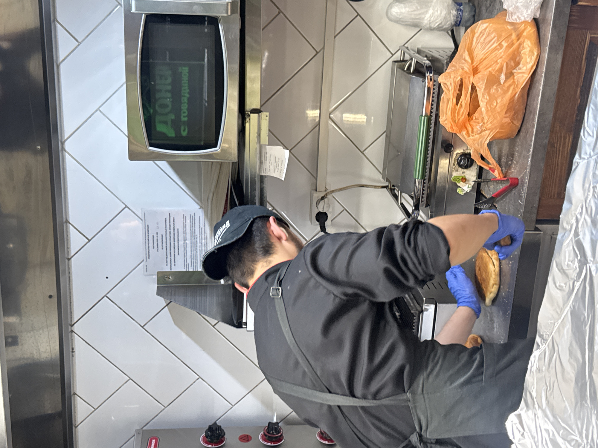 A man in a black uniform and blue gloves is preparing food in a kitchen. The man is standing in front of a grill or sandwich press. He appears to be placing or adjusting a bread product, possibly a sandwich or pita, into the press. A crumpled orange plastic bag sits near the grill, and rolls of aluminum foil are visible to the right. A microwave with a sign reading "Донер с говядиной" (Doner with Beef) in Russian is positioned behind him and to the left, attached to a wall with tile. A document is taped to the wall above a row of knobs. The location is consistent with a commercial kitchen setting in Bugry, Russia.