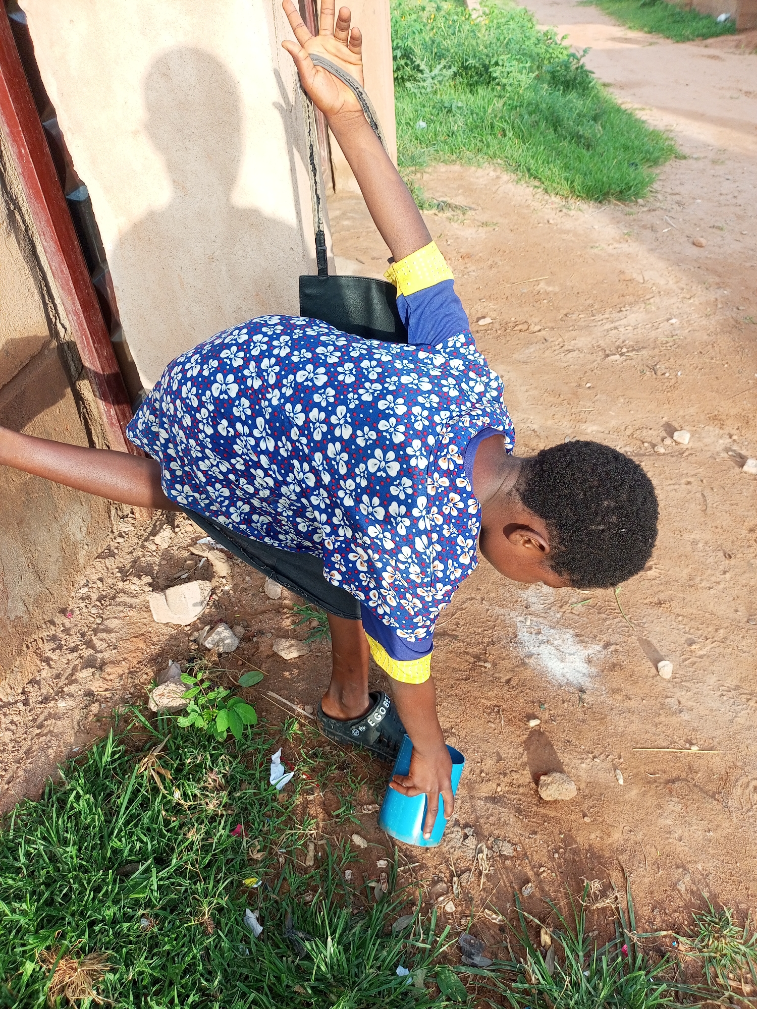 An overhead, high-angle shot shows a young person from the shoulders down, leaning forward and reaching with their right hand for a blue plastic cup on the ground. The individual is wearing a blue shirt with white and red floral patterns and yellow cuffs, and black pants. Their left arm is extended upwards, leaning against a rough, textured wall. The ground is a mix of dry, reddish-brown dirt, patches of green grass, and scattered stones and debris. In the background, a dirt path winds past a grassy area. The scene appears to be outdoors in a somewhat unkempt area, possibly a street or yard in Tempure, Nigeria. Sunlight casts a distinct shadow of the person on the wall.