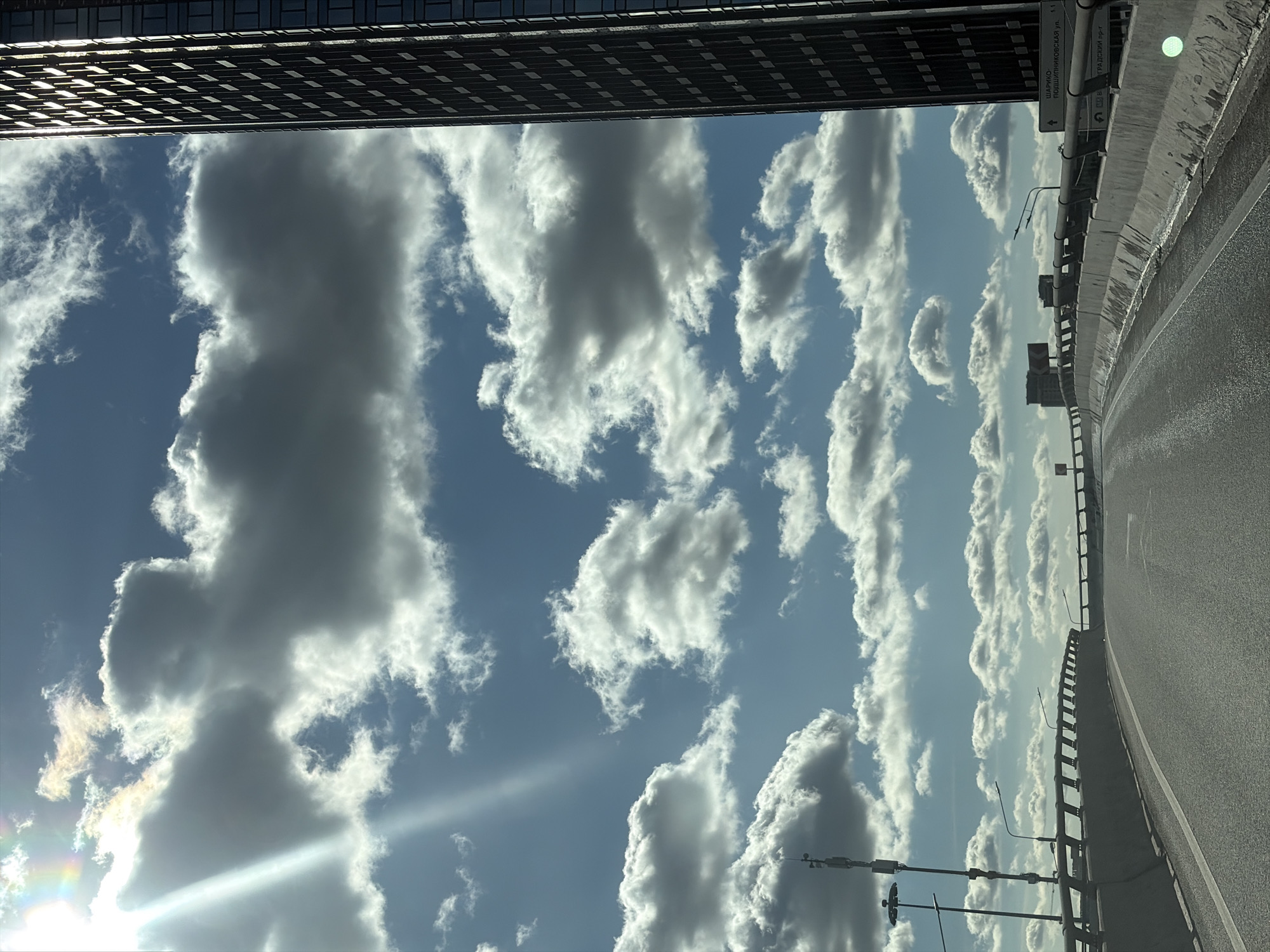 The image captures a view from a car traveling on an elevated highway in Moscow, Russia, likely on a day with a partly cloudy sky. To the right, a tall, modern skyscraper with a dark facade and repeating rectangular windows dominates the skyline. The highway itself is paved and has concrete barriers with metal railings along its edge. Streetlights and communication towers are visible along the highway. The sky is a vibrant blue with scattered white and gray cumulus clouds, and a bright sunbeam with a hint of a rainbow breaks through from the upper left. The overall impression is one of urban infrastructure against a dynamic sky. Signs are visible on the highway, one of which reads "ШАРИКО ПОДШИПНИКОВСКАЯ ул." which translates to "Sharikopodshipnikovskaya street."
