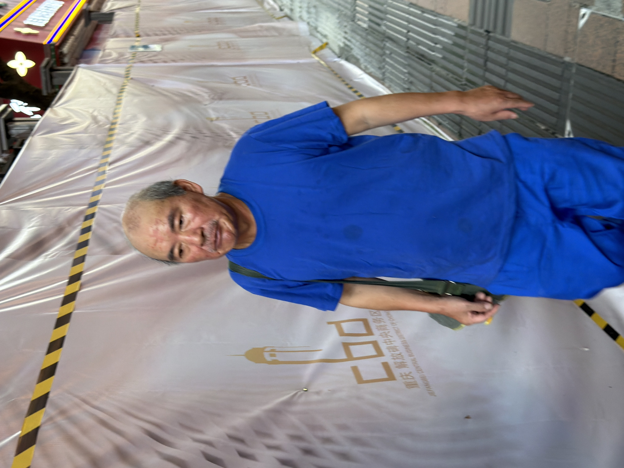 A middle-aged East Asian man with a bald head and a mustache, wearing a bright blue t-shirt and matching blue pants, walks past a large white banner with a gold logo. The banner features Chinese characters that read "重庆解放碑中央商务区" (Chóngqìng Jiěfàngbēi Zhōngyāng Shāngwùqū) and English text "JIEFANGBEI CENTRAL BUSINESS DISTRICT OF CHONGQING". The man has a green strap from a bag visible across his shoulder and is holding his right hand near his side. He appears to be walking on a sidewalk with metal grating. Yellow and black caution tape is visible running horizontally across the banner and along the top of a barrier to the right. The background suggests an urban environment with some construction or renovation in progress, indicated by the draped banner.