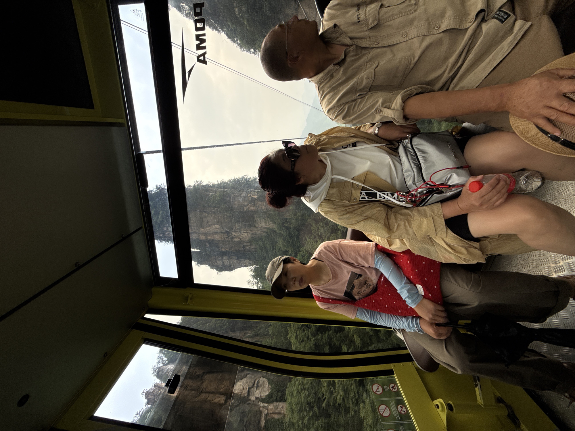 Inside a Zhonghu, China cable car, three people are seated. An elderly man in a tan shirt sits next to a woman in a yellow jacket holding a water bottle and a silver bag. Another woman wearing a pink shirt and a red bag is also present. They are overlooking the mountainous landscape through the windows.