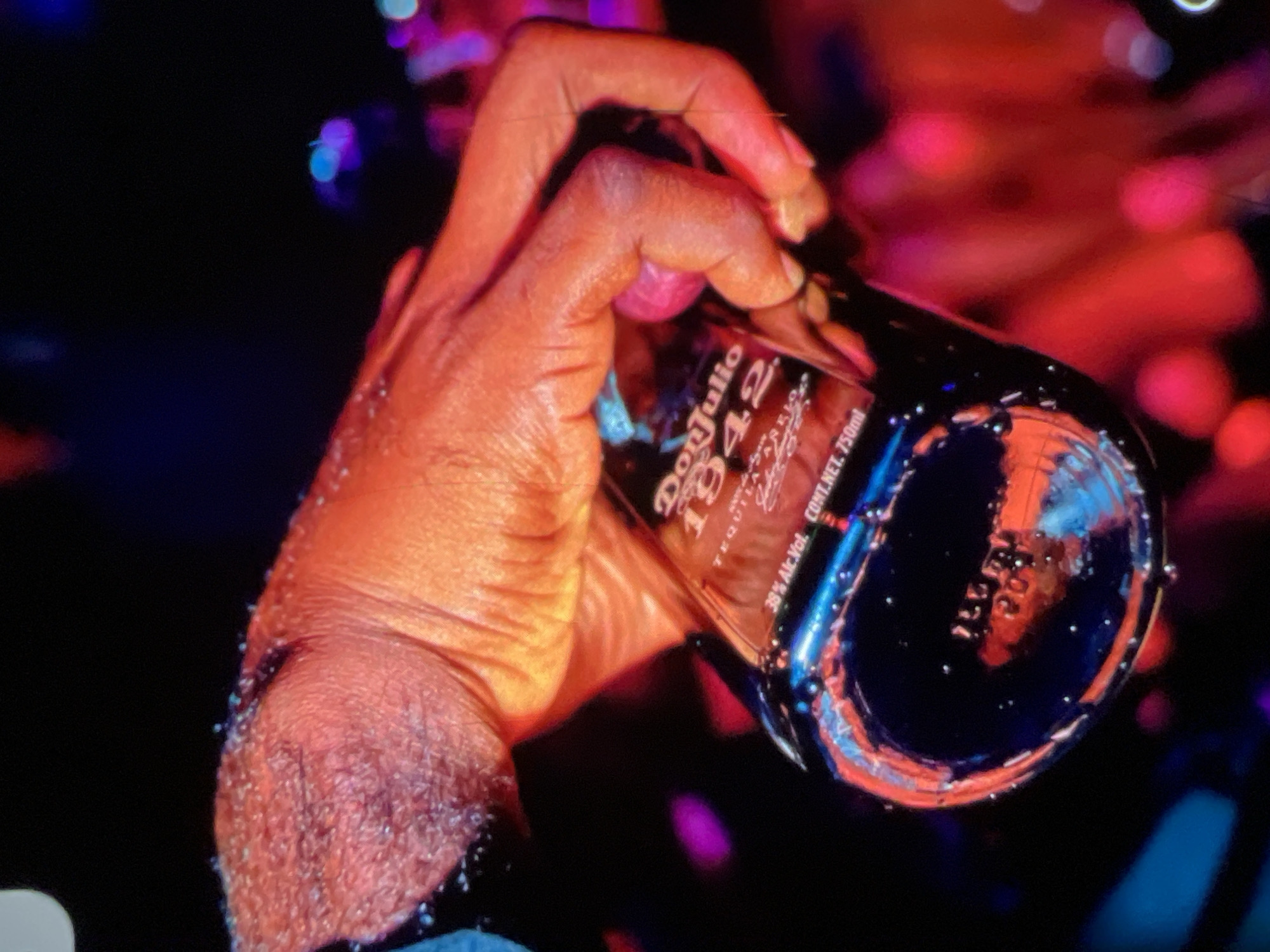 A close-up shot focuses on a person's hand holding a bottle of Don Julio 1942 tequila. The hand, with visible pores and some light glitter, is illuminated by warm orange and red lights, suggesting a vibrant, possibly nightlife, setting. The bottle is partially submerged in a dark liquid, and the label is clearly visible, indicating the brand and "1942" as a prominent detail. Small droplets of liquid cling to the outside of the bottle. The background is dark with blurred, colorful bokeh lights in shades of pink, blue, and purple, further enhancing the atmosphere of a party or event. The image does not contain any information that allows for the determination of its location in Lagos, Nigeria.