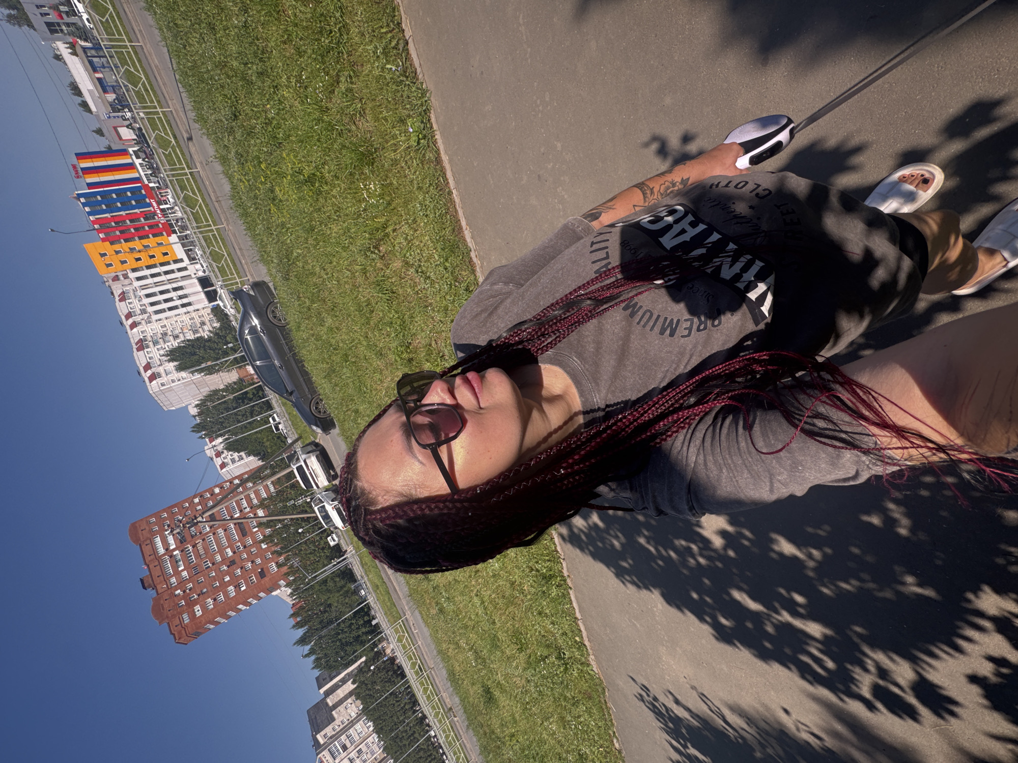 A woman with braided hair and sunglasses is taking a selfie outdoors in Tolyatti, Russia, on a sunny day. She is wearing a grey t-shirt with graphics that read "PREMIUM" and "IMAGINE" and white slide sandals. She is holding a retractable dog leash in her right hand. In the background, there are several multi-story buildings, some with colorful facades, a grassy area, a road with cars, and a clear blue sky. One building in the background is a tall, red brick structure with many windows. Another building has vibrant colored vertical stripes of yellow, blue, and red. The scene suggests an urban environment.